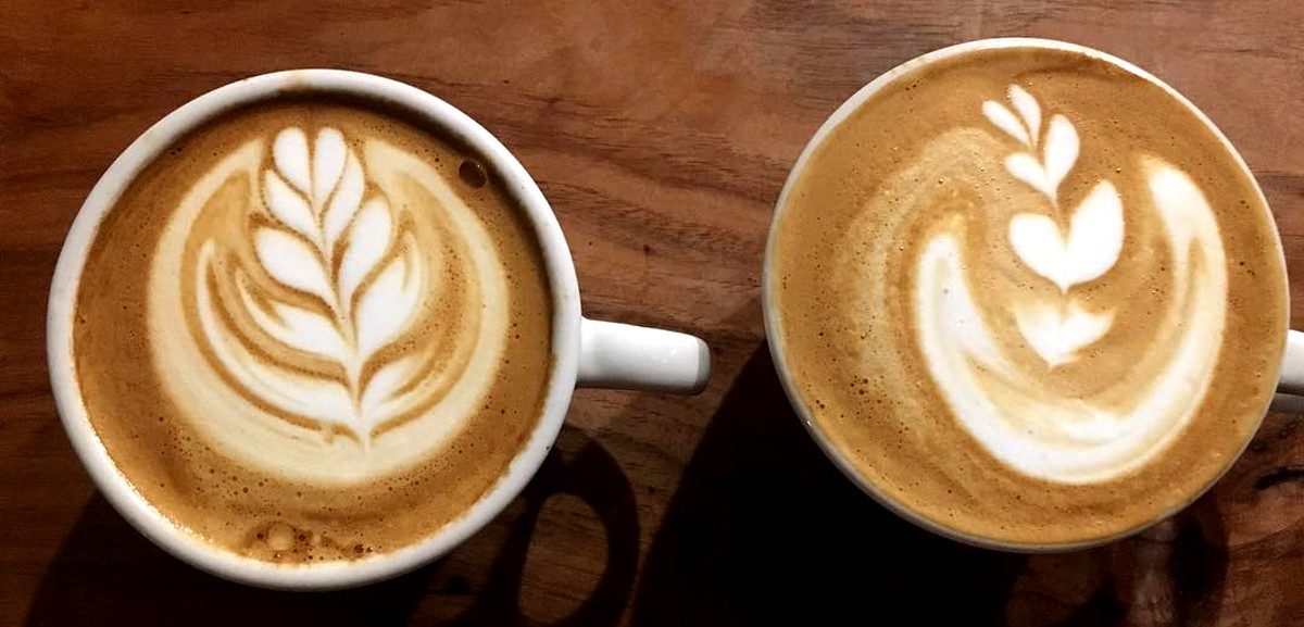 Two cups of specialty coffee with latte art rest on a wooden surface. The left cup shows a leaf design, while the right displays a similar, slightly asymmetrical leaf—both showcasing the craft of skilled coffee roasters.