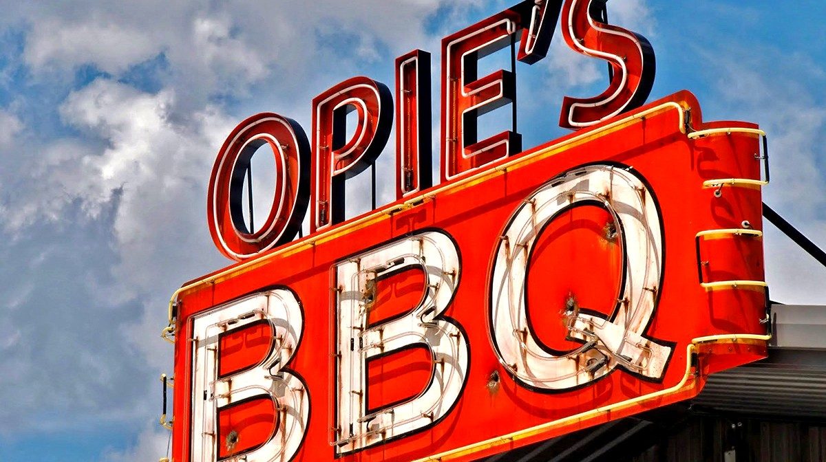 A vintage-style neon sign reads OPIES BBQ in bold red and white letters against a cloudy sky, advertising Opie's Barbecue, a classic Texas barbecue restaurant.