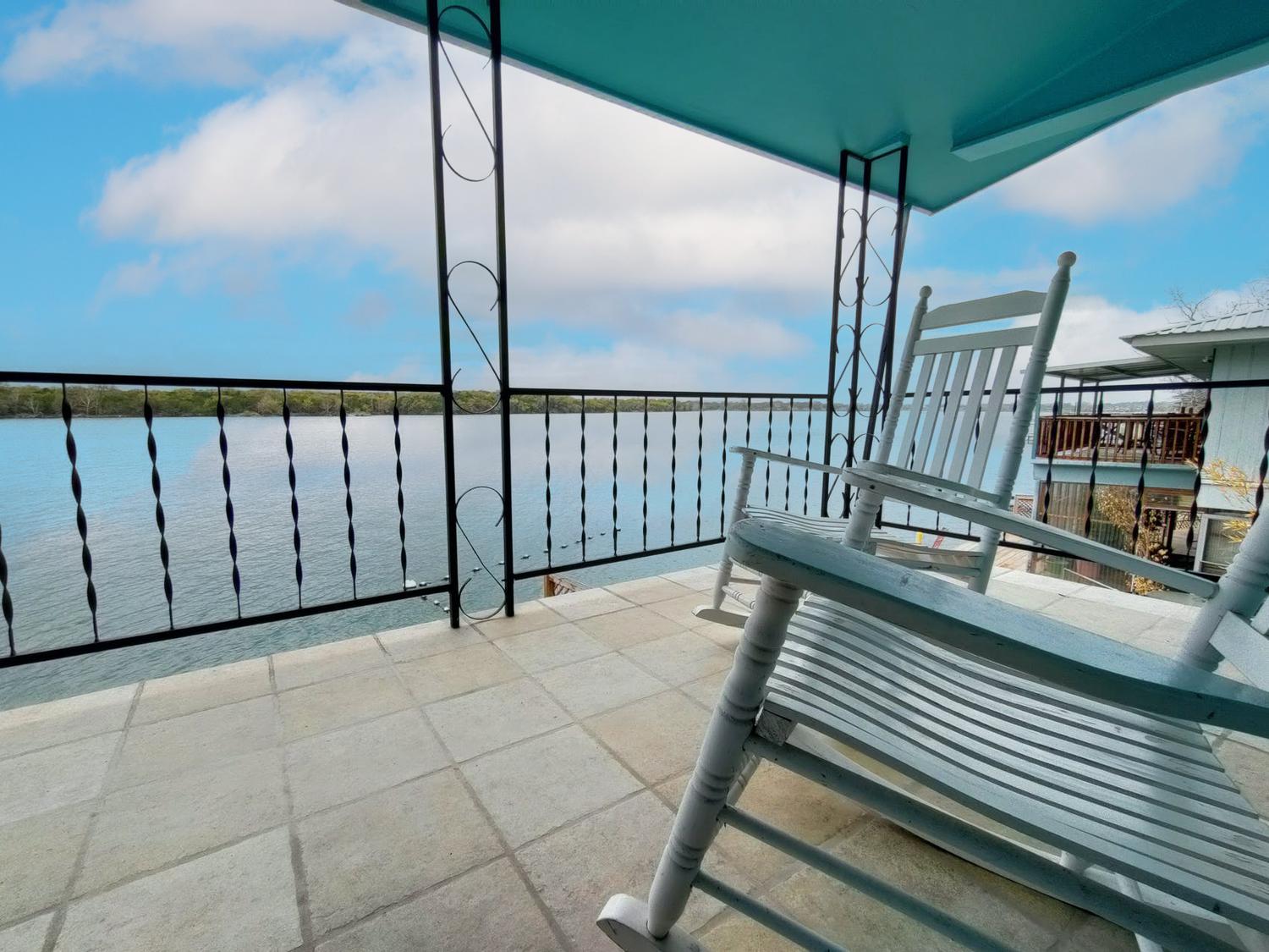 A white rocking chair sits on the tiled balcony at The Lookout, overlooking a calm lake with a black metal railing, blue ceiling above, and views of trees across the water—an inviting spot reminiscent of LBJ’s peaceful retreats.
