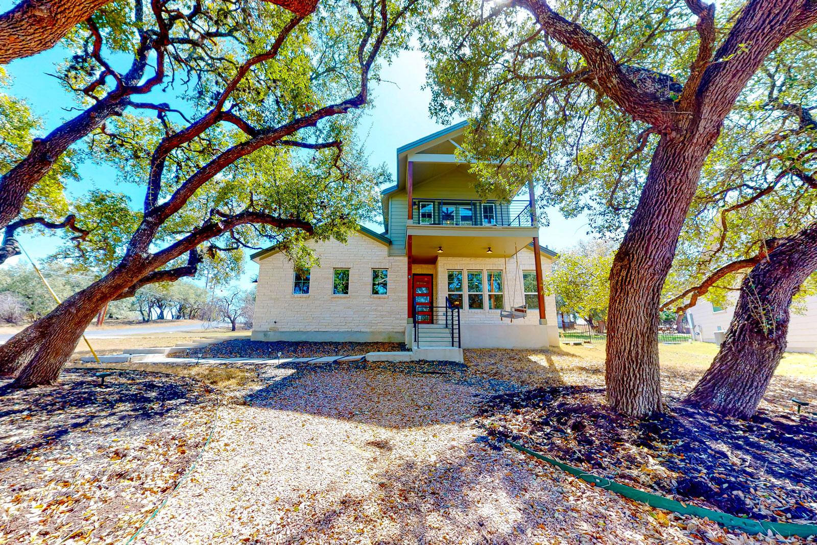 A luxury two-story farmhouse with a front porch and balcony, light-colored brick exterior, red door, surrounded by large oak trees and a leaf-covered yard on a sunny day.