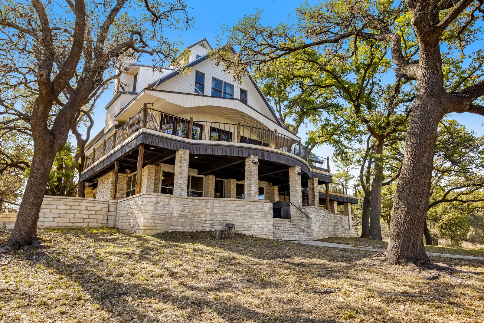 Large, three-story house with stone and wood exterior, wraparound porch, and balcony overlooking Lake Travis, surrounded by mature trees on a sloped yard under a clear blue sky.