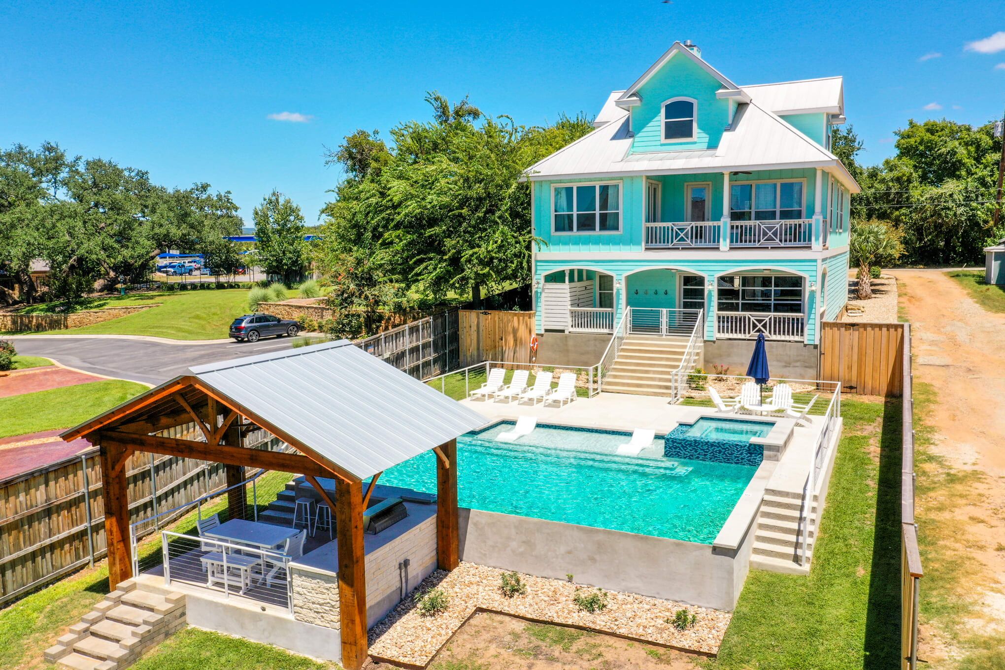 A two-story turquoise house with white trim overlooks a large backyard pool. The pool has a covered pavilion with seating and a grill, surrounded by lounge chairs and a wooden fence, on a sunny day.