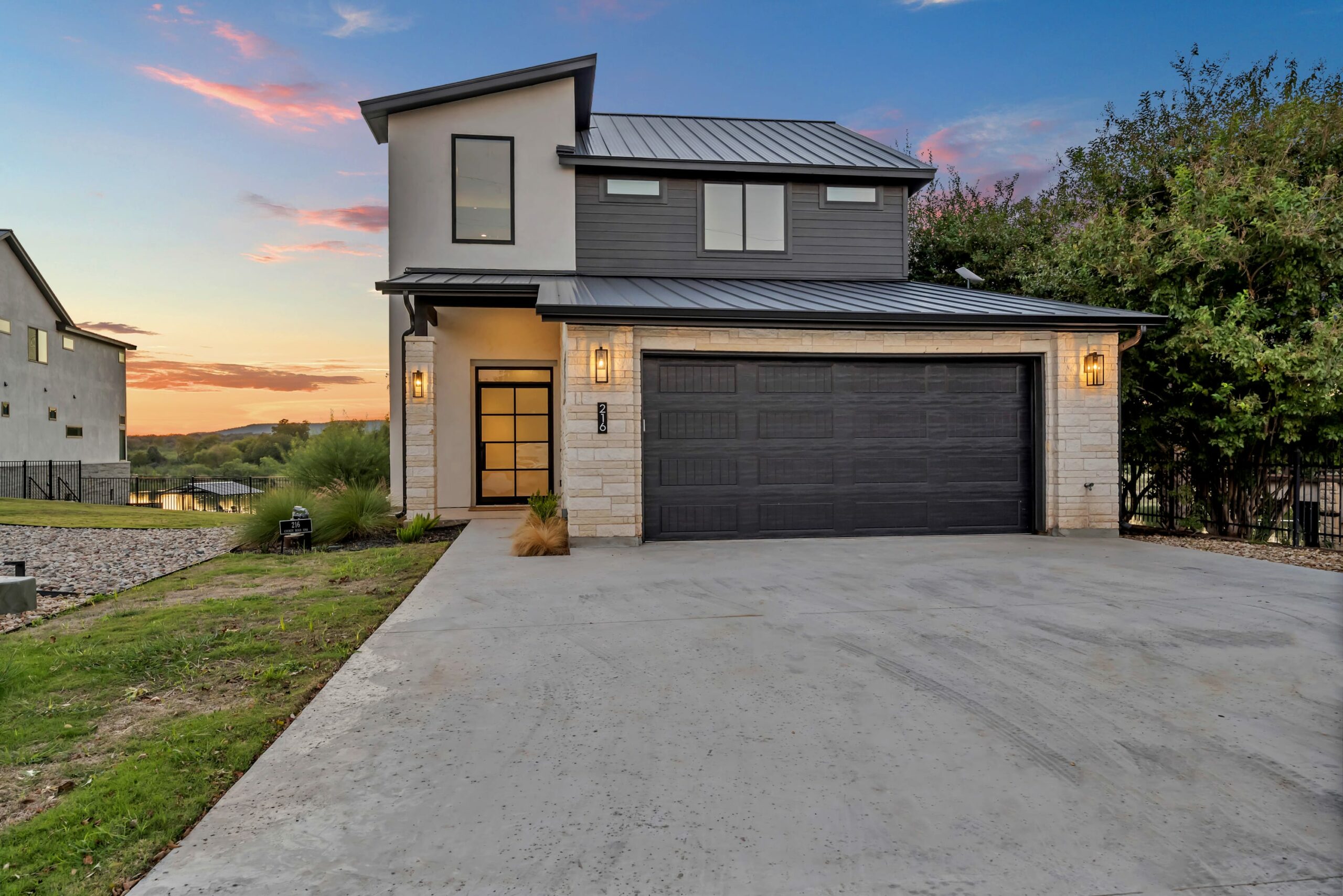 Modern two-story house with dark roofing, stone and wood exterior, large windows, and a double garage; driveway leading to the entrance of this LBJ Bliss Retreat, with sunset sky and lush greenery in the background.