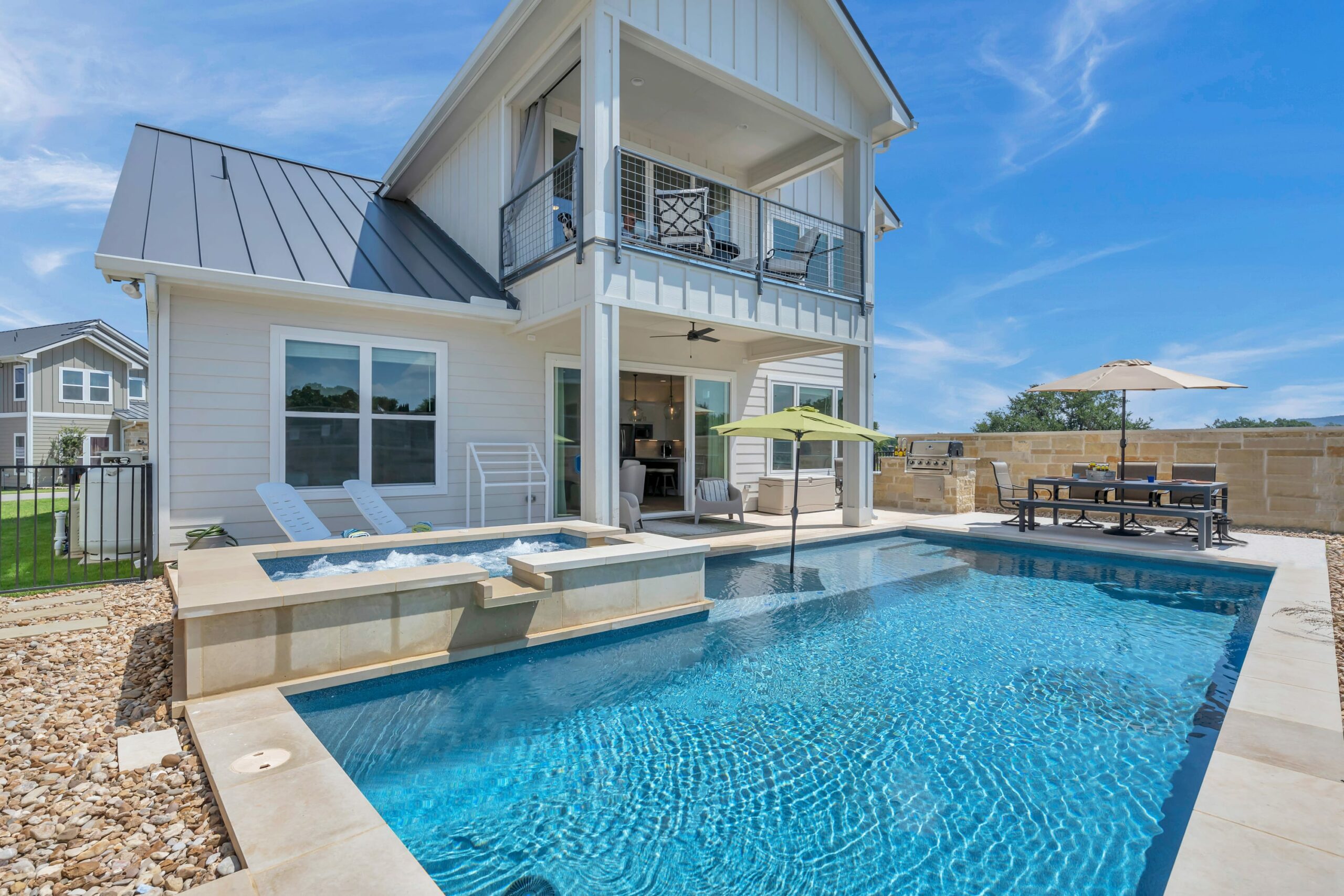 A modern two-story house, Heavenly Haven, features a backyard pool, jacuzzi, lounge chairs, and a shaded outdoor dining area. The weather is sunny with a clear blue sky.
