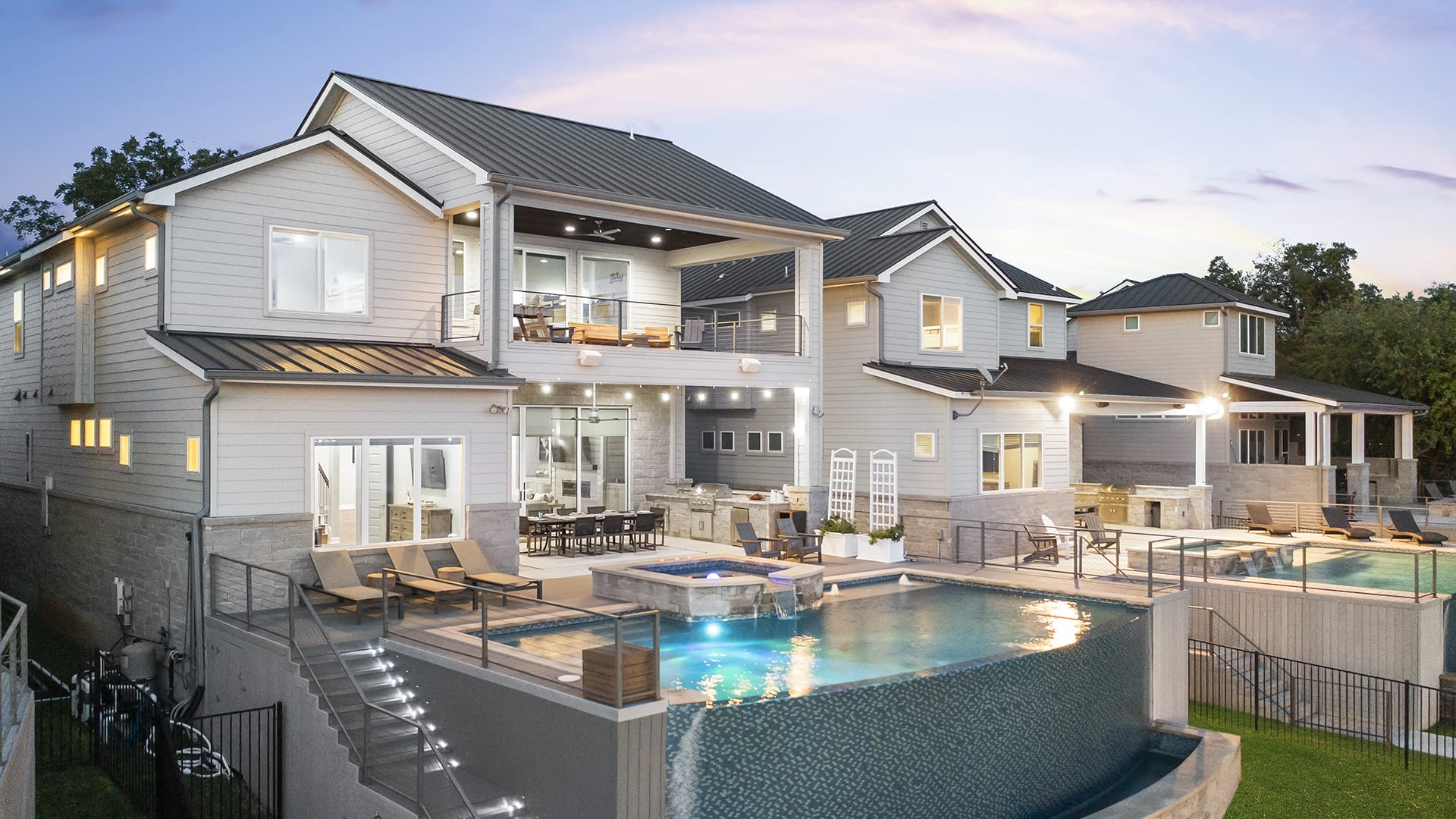 Modern two-story Bud Shores Sanctuary with large windows, covered balconies, and a sleek infinity pool with hot tub. Outdoor seating areas and landscape lighting create a luxurious, inviting atmosphere at dusk.