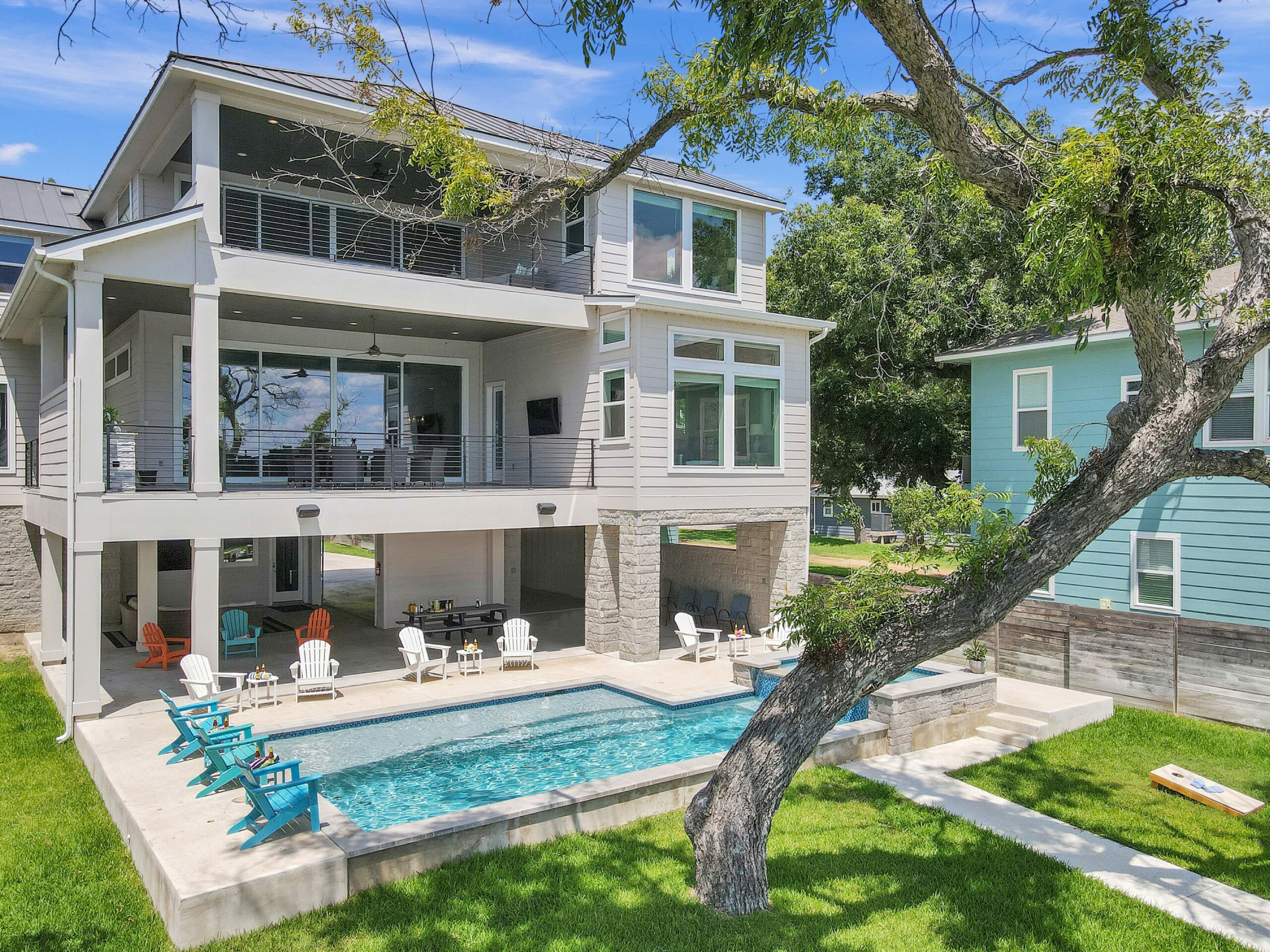 Modern two-story house with large windows, covered balcony, and a backyard oasis—Rolling Waves Haven boasts a sparkling pool, lounge chairs, lush green lawn, and a graceful tree reaching forward in the foreground.
