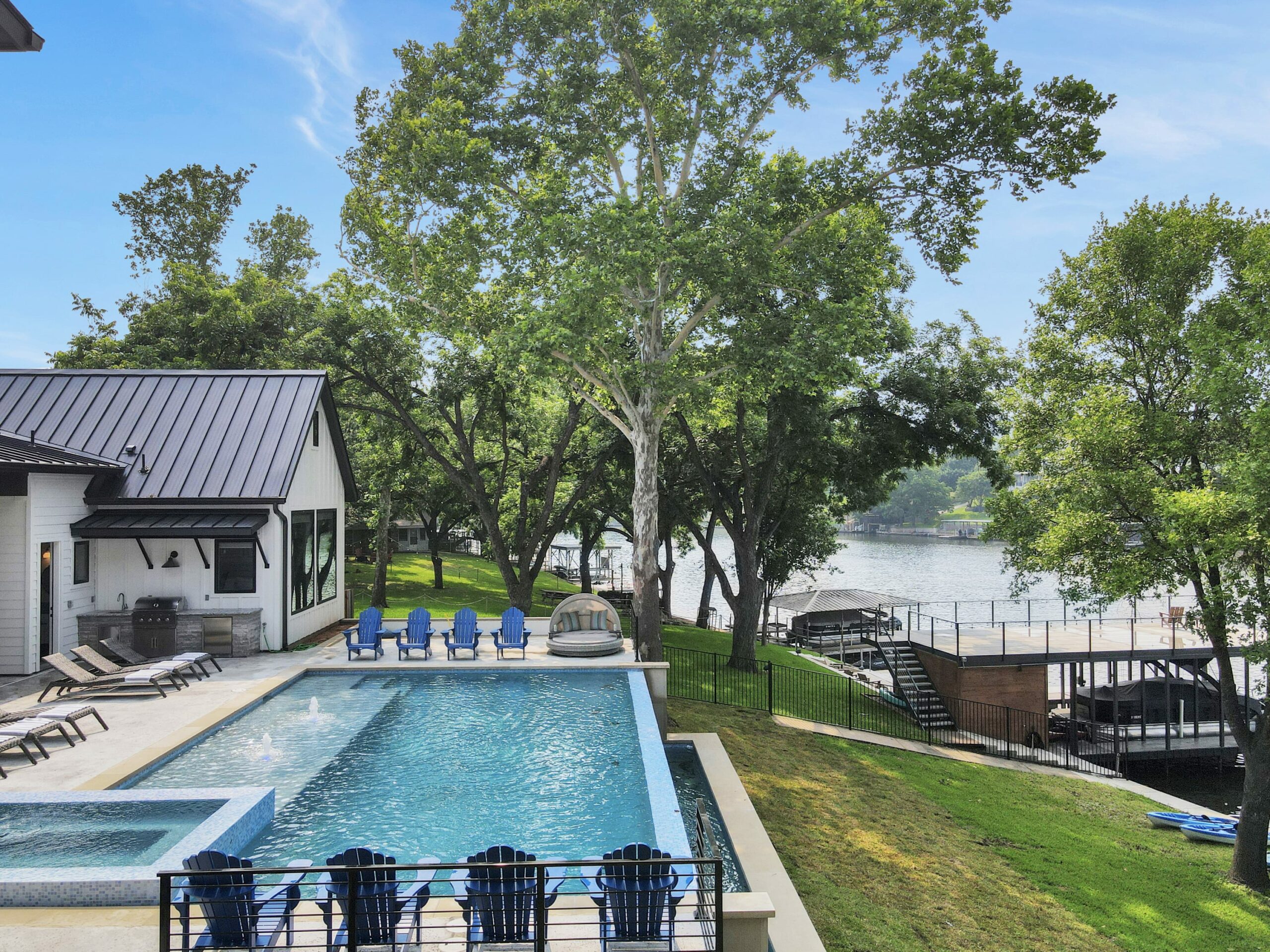 Modern lakeside home with swimming pool, lounge chairs, and shaded patio area. Lush green lawn leads to a dock on Lake LBJ, surrounded by trees and clear blue sky—your perfect Palm Paradise vacation rental.