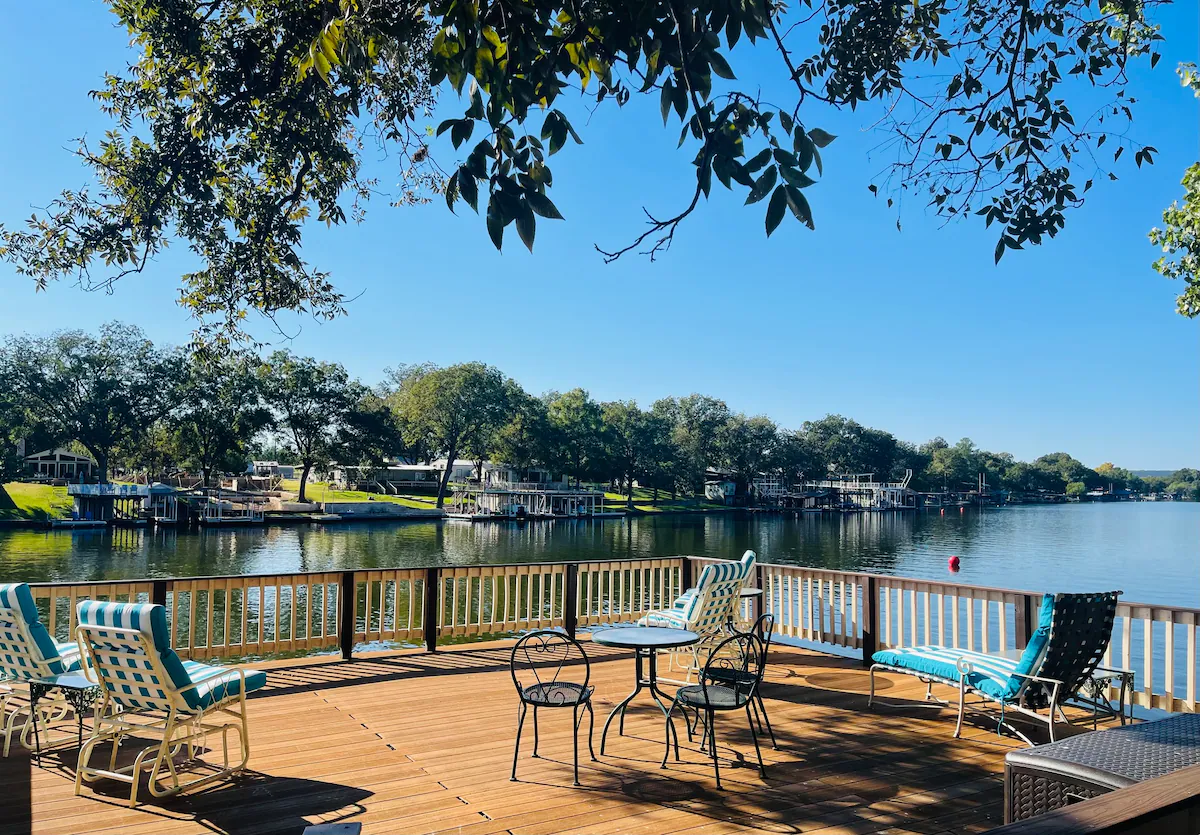 A spacious wooden deck at 1100 Lakeside Drive, with lounge chairs and a table, overlooks a calm lake surrounded by trees and waterfront houses beneath a clear blue sky.
