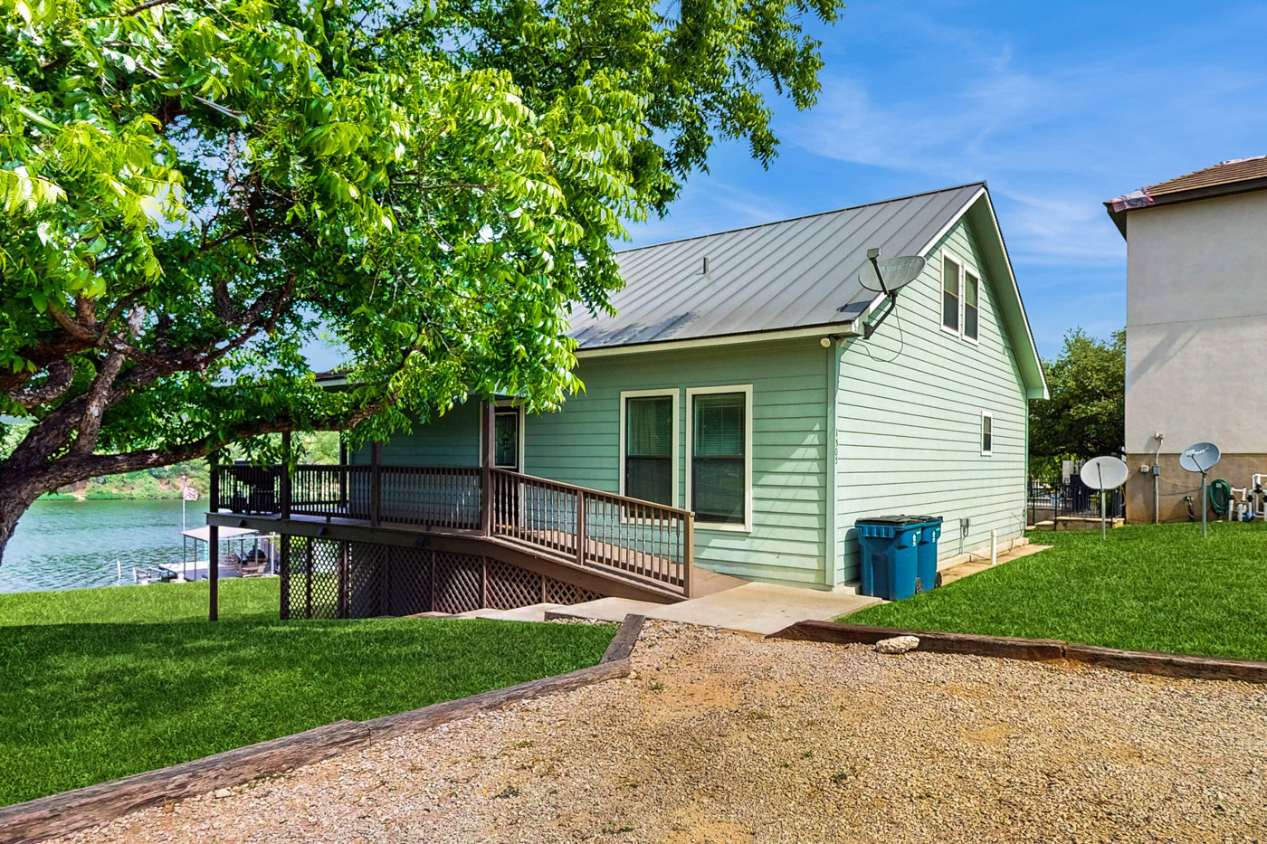 A light green RUFF Lakehouse with a metal roof sits near Lake LBJ. A wooden ramp leads to the entrance. Lush grass, a large tree, and blue bins are visible; a dock can be seen on the water in the background—perfect for your vacation rental.