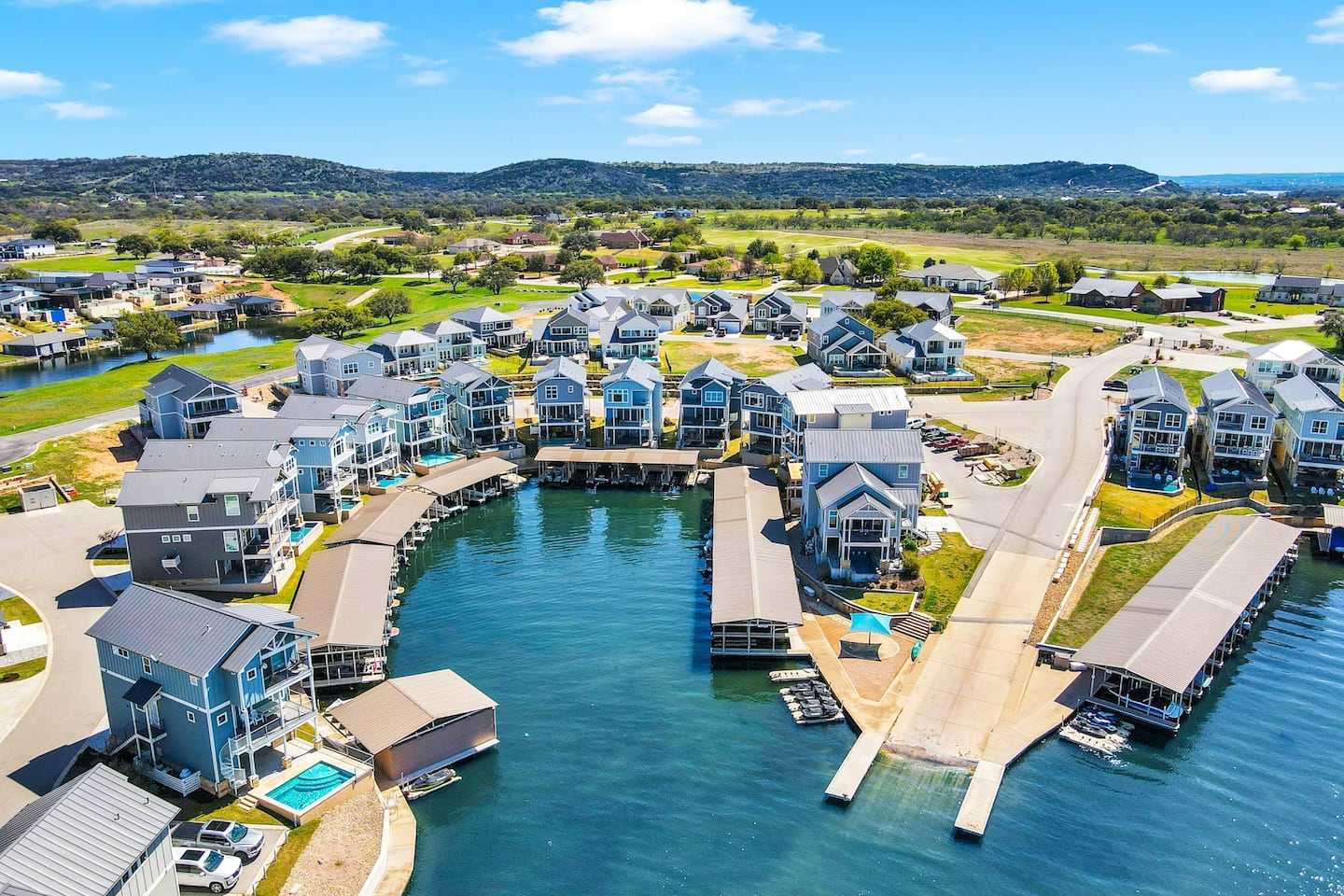 Aerial view of Blue Water Bluffs, a waterfront neighborhood with blue and white houses, private docks, and boat ramps curving around a sheltered cove, surrounded by green hills under a bright, clear sky.