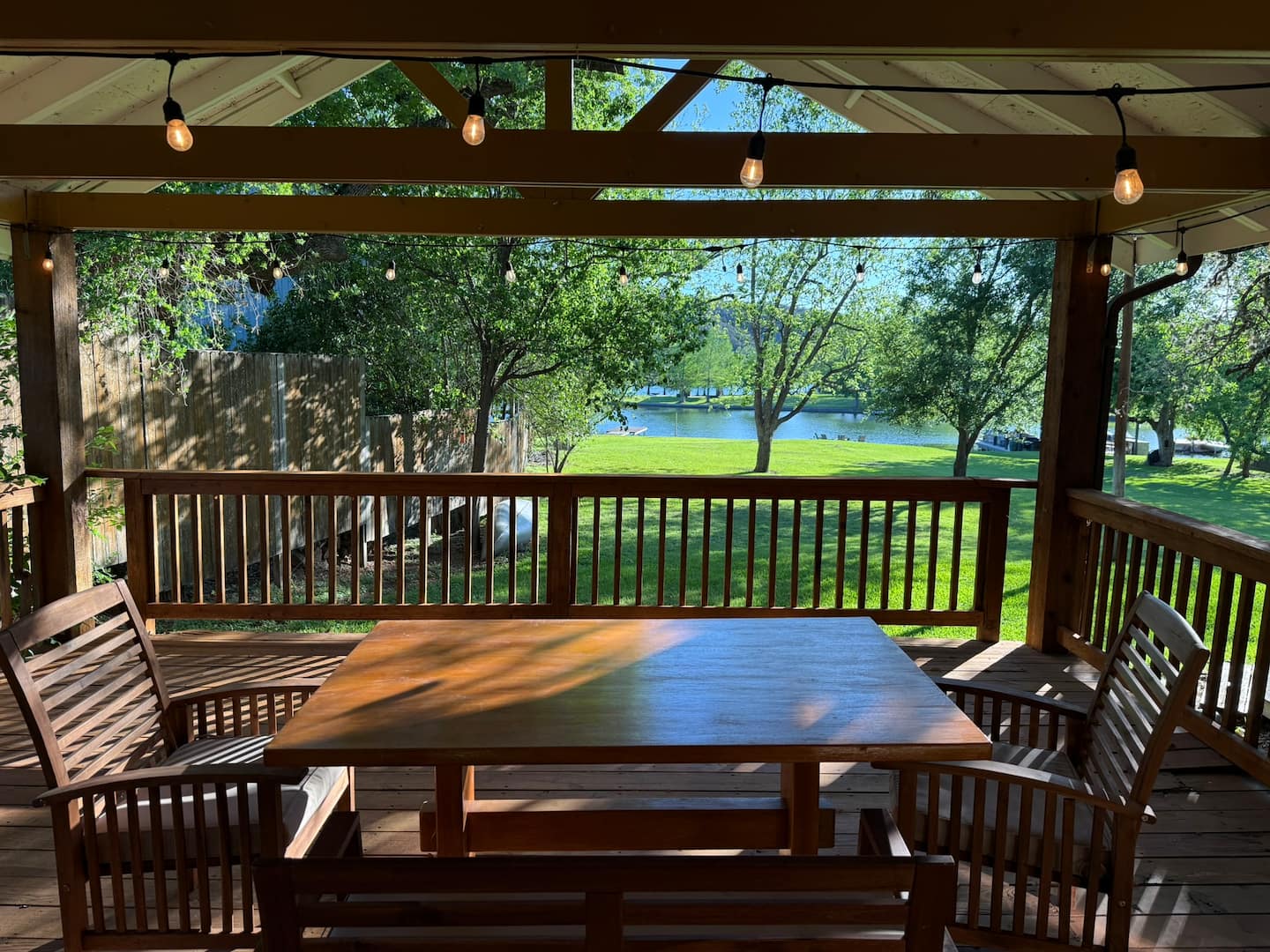 A wooden patio with a table and chairs sits under a covered roof with string lights. The deck at the historic Thompson-Kinney House overlooks a grassy yard, trees, and a river on a sunny day.