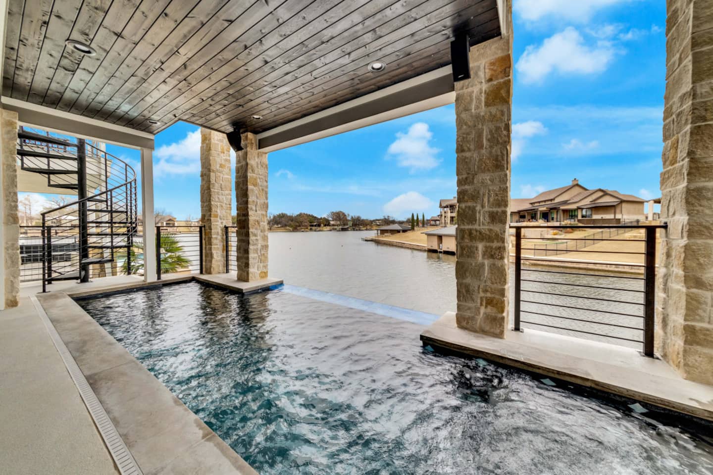Covered patio with LONESTAR CABANA vibes, infinity-edge pool, stone pillars, and a view of a calm lake bordered by houses. A spiral staircase is visible on the left beneath a bright sky with scattered clouds.