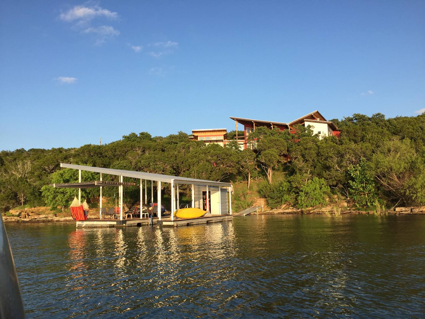A modern house sits on a tree-covered hill above a lake. Below, a small dock with a slanted roof holds a kayak, dock chairs, and a swing—an ideal sunset escape with sunlight reflecting on the calm water.