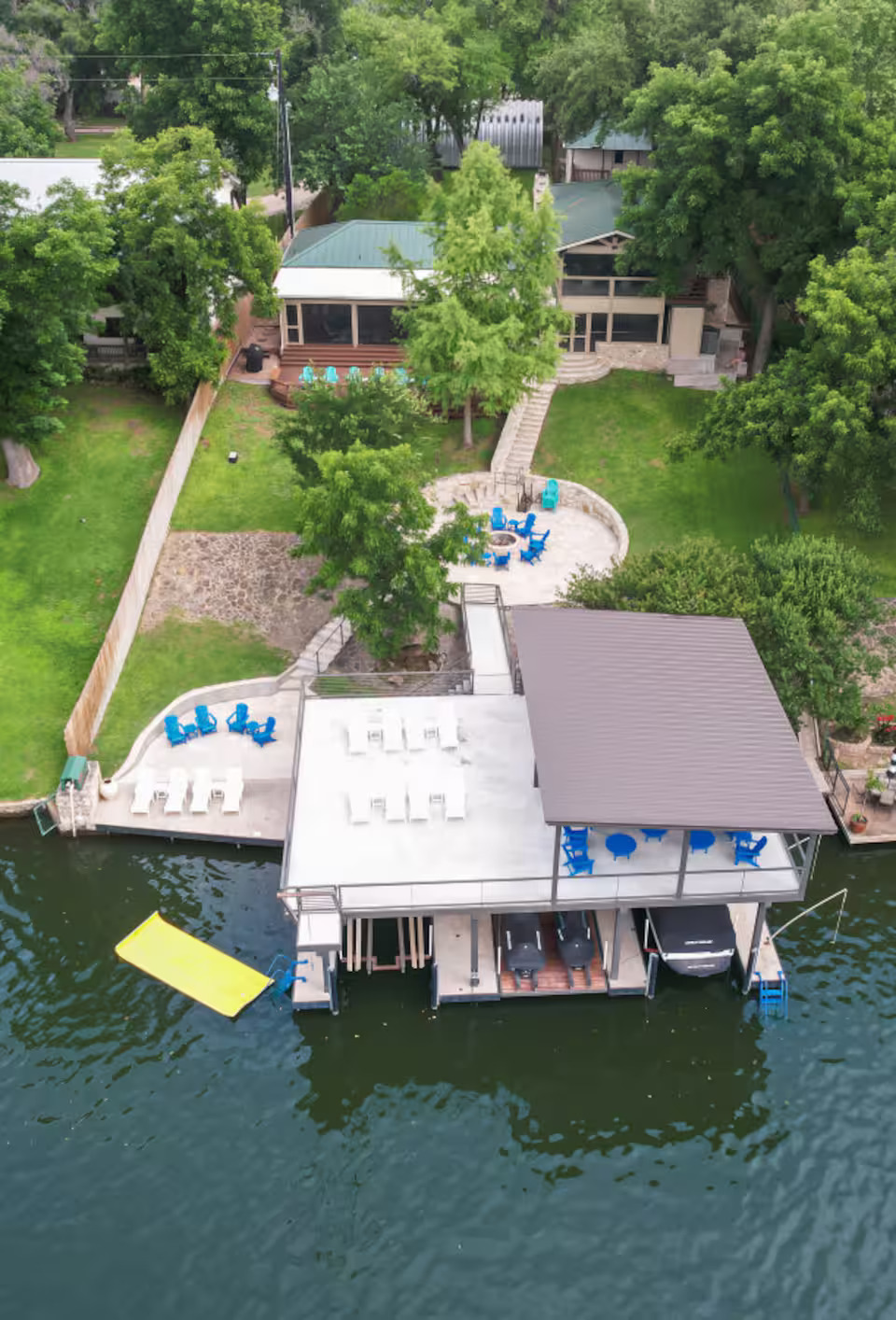 Aerial view of a lakeside Gathering Place on LBJ, featuring a two-level dock, blue and white seating areas, a yellow floating mat in the water, and a green lawn with trees leading to the house in the background—ideal as an event venue.