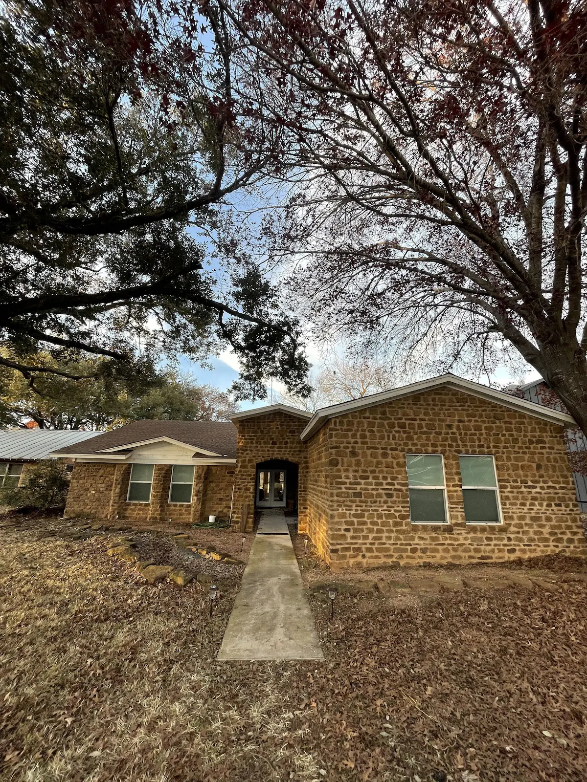 Single-story brick house with large front windows, a central entrance, and a concrete walkway—this tranquil getaway is surrounded by leafless trees and dry grass under a partly cloudy sky near Lake LBJ.