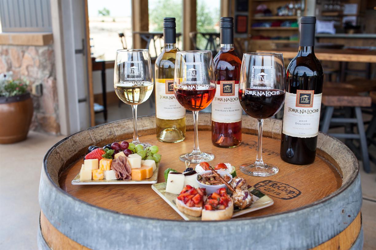 A wine barrel holds three glasses of wine, four bottles of assorted wine, and two plates with assorted cheeses, meats, fruit, and bruschetta, set in a rustic winery tasting area.