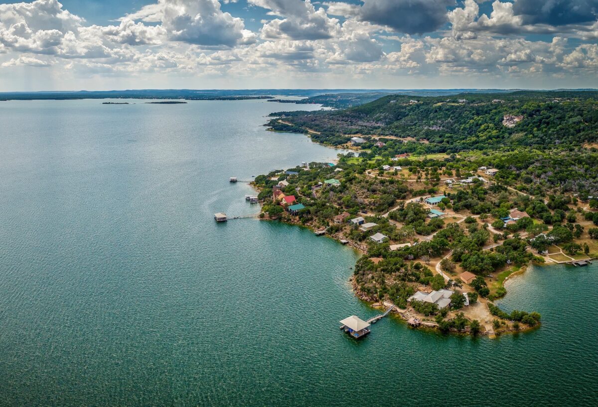 Aerial view of a lake with calm blue-green water, docks, and houses scattered along a lush, tree-covered shoreline under a sky with scattered clouds.