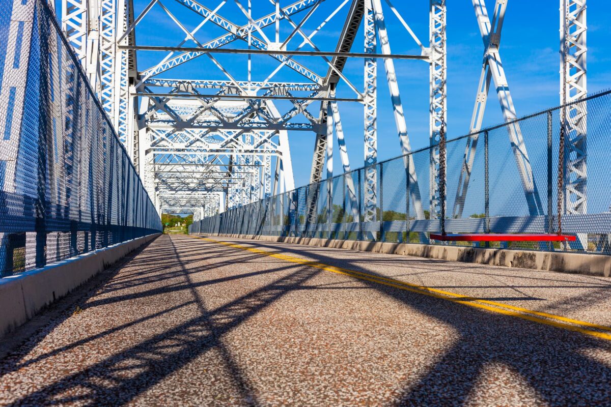 View of a steel truss bridge from the road surface, with a yellow center line, metal railings, and blue sky above. Shadows from the bridge structure are cast onto the pavement.