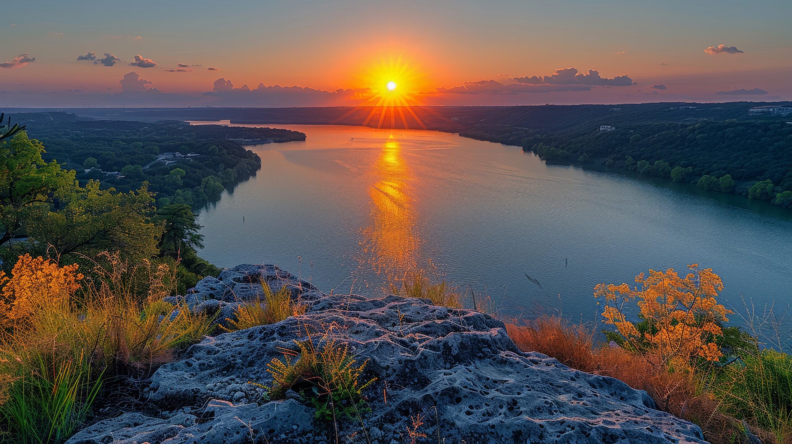 Sunset over a wide river viewed from a rocky, grassy cliff. The sun casts a golden reflection on the water, surrounded by lush green trees and distant hills under a clear sky with a few clouds.