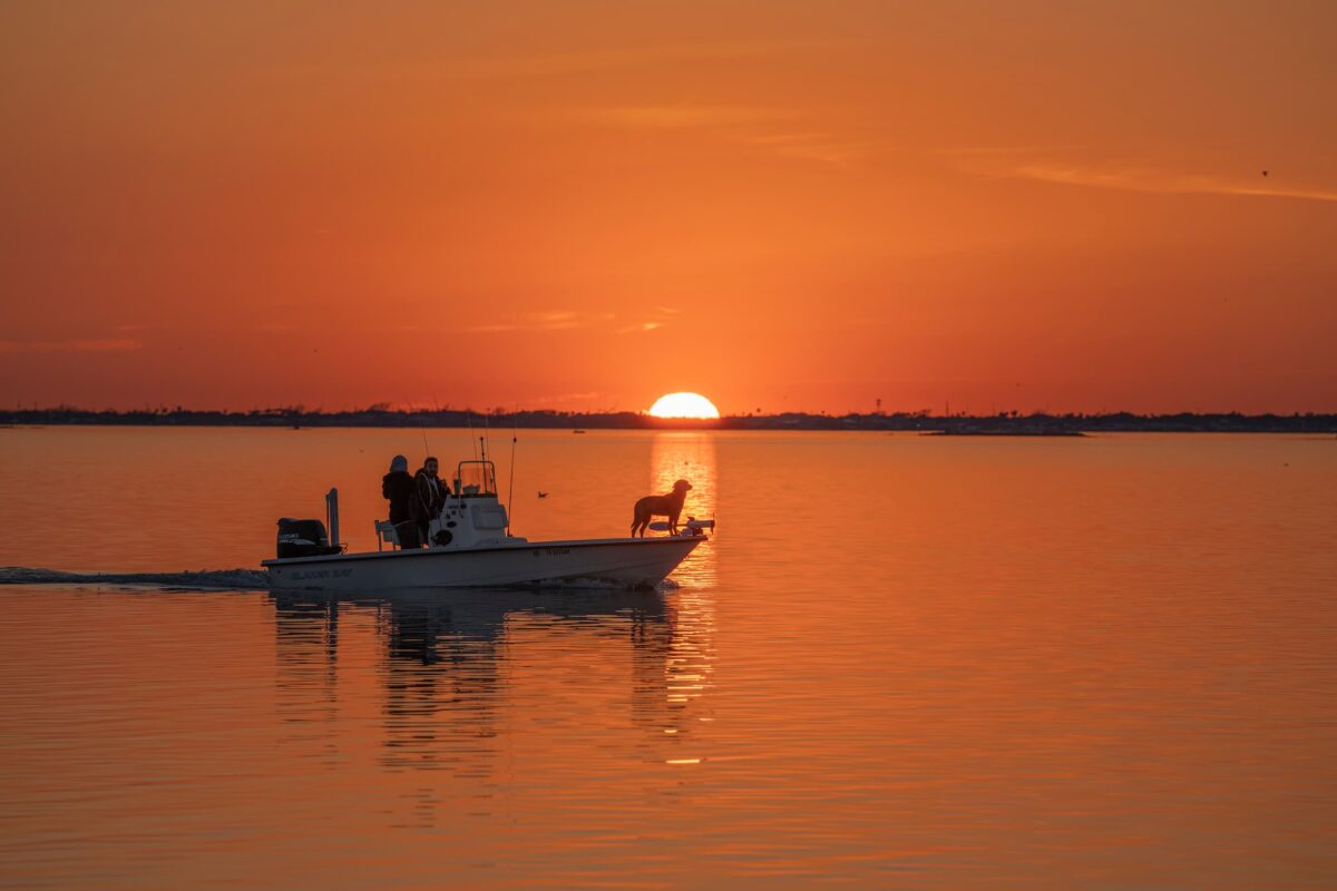A small motorboat with two people and a dog glides across calm water on Lake Buchanan at sunset, the orange sun low on the horizon and its reflection shimmering on the surface.