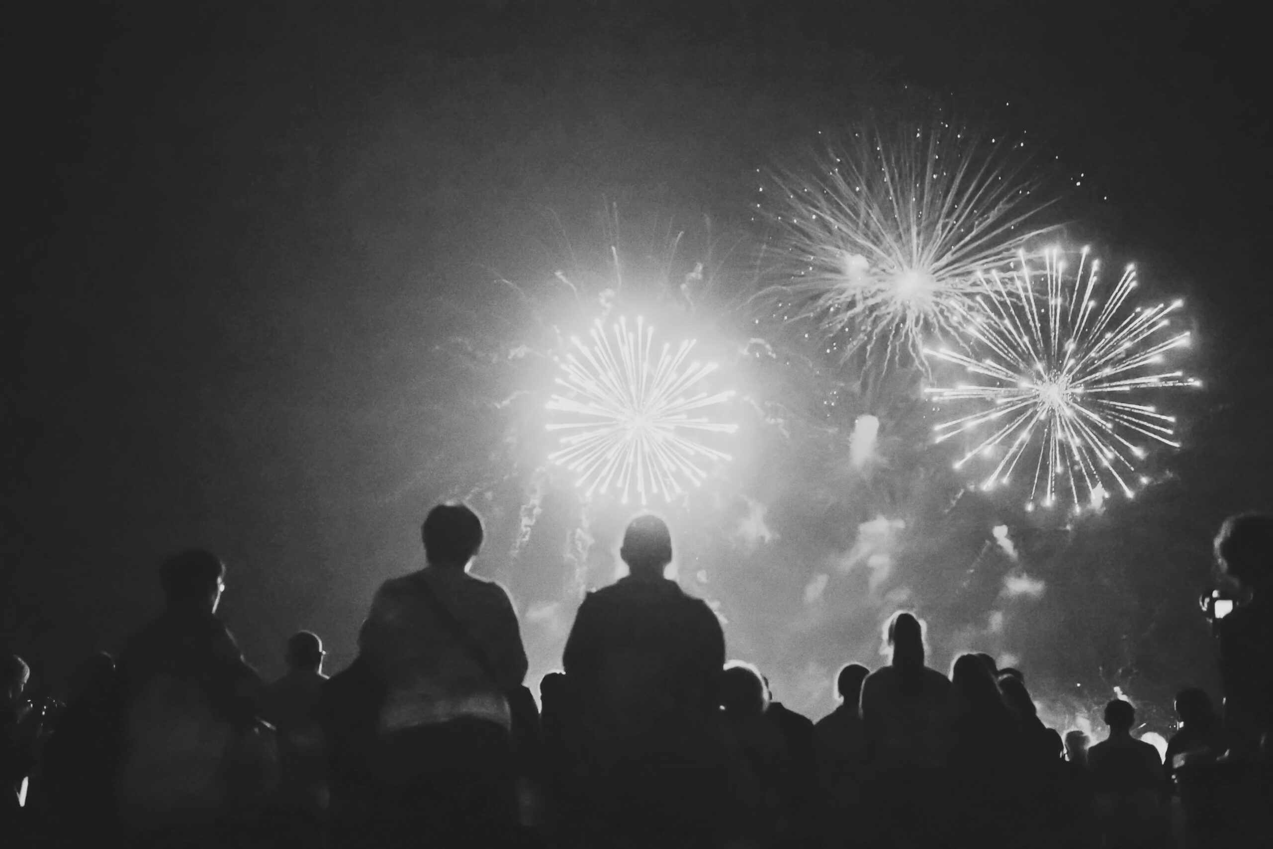 A crowd of people watches fireworks exploding in the night sky. The scene is in black and white, with bright fireworks illuminating the silhouettes of the spectators.
