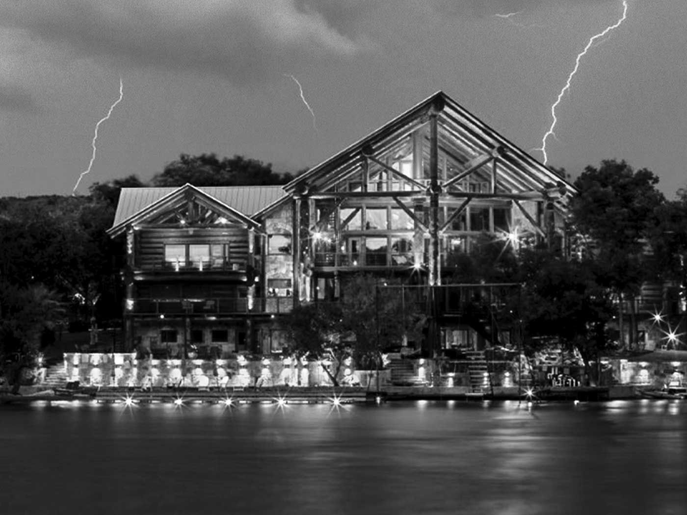 A large, modern log cabin with glass-front windows sits by the water at night—an ideal spot for those seeking where to stay. Bright lights illuminate the house as lightning bolts flash across the cloudy, black and white sky.