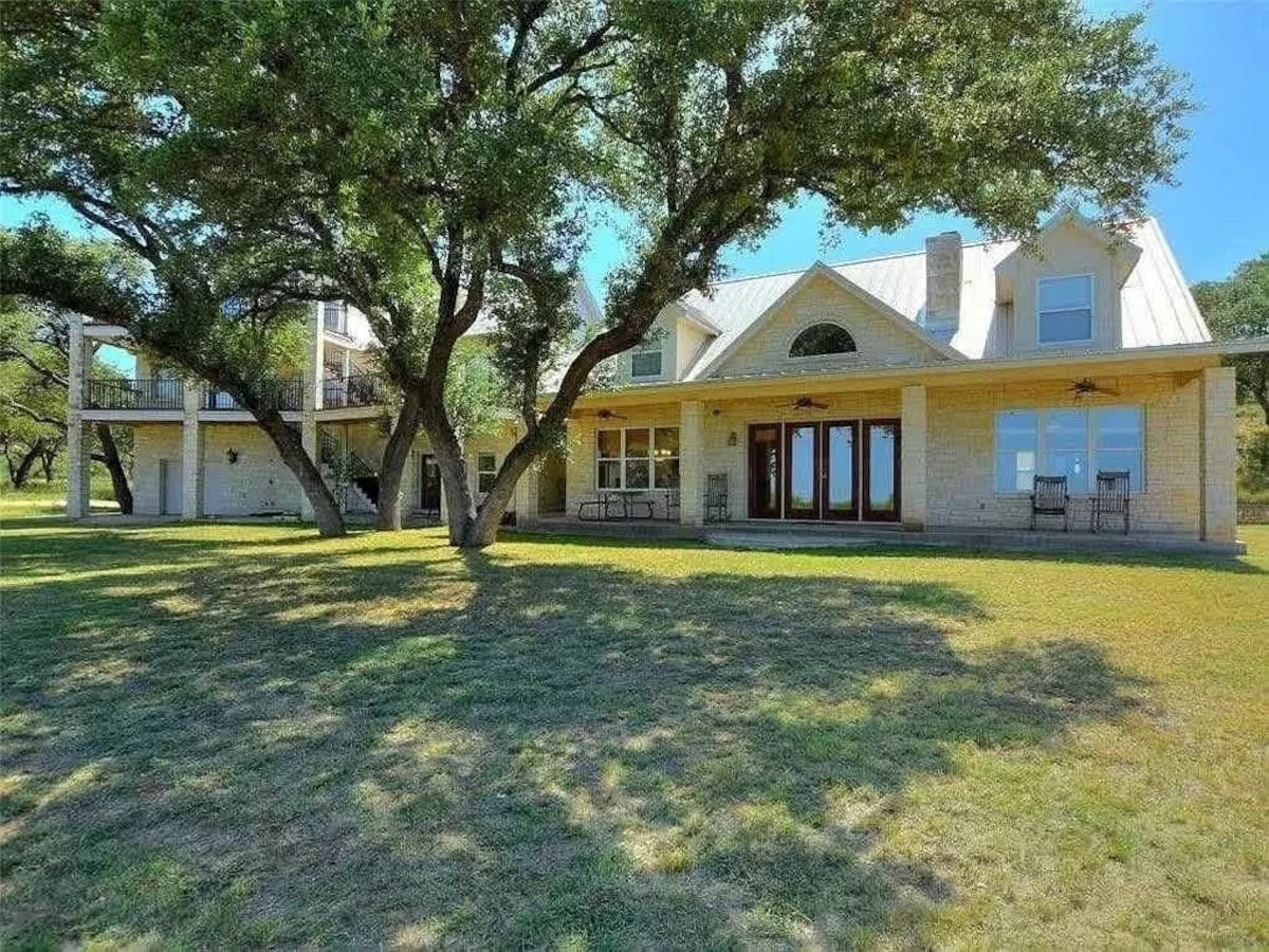 Two-story stone house with a metal roof, large windows, covered porch, and balcony sits amid the TX Kelly Emeralds, surrounded by green grass and shaded by tall trees on a sunny day.