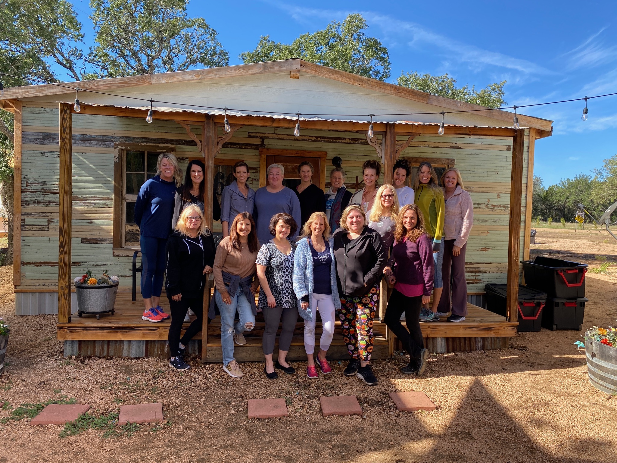 A group of 18 women pose and smile together on the wooden porch of a rustic Texas Ranch Retreat, surrounded by trees under a blue sky. Some are standing, while others sit or kneel in front.