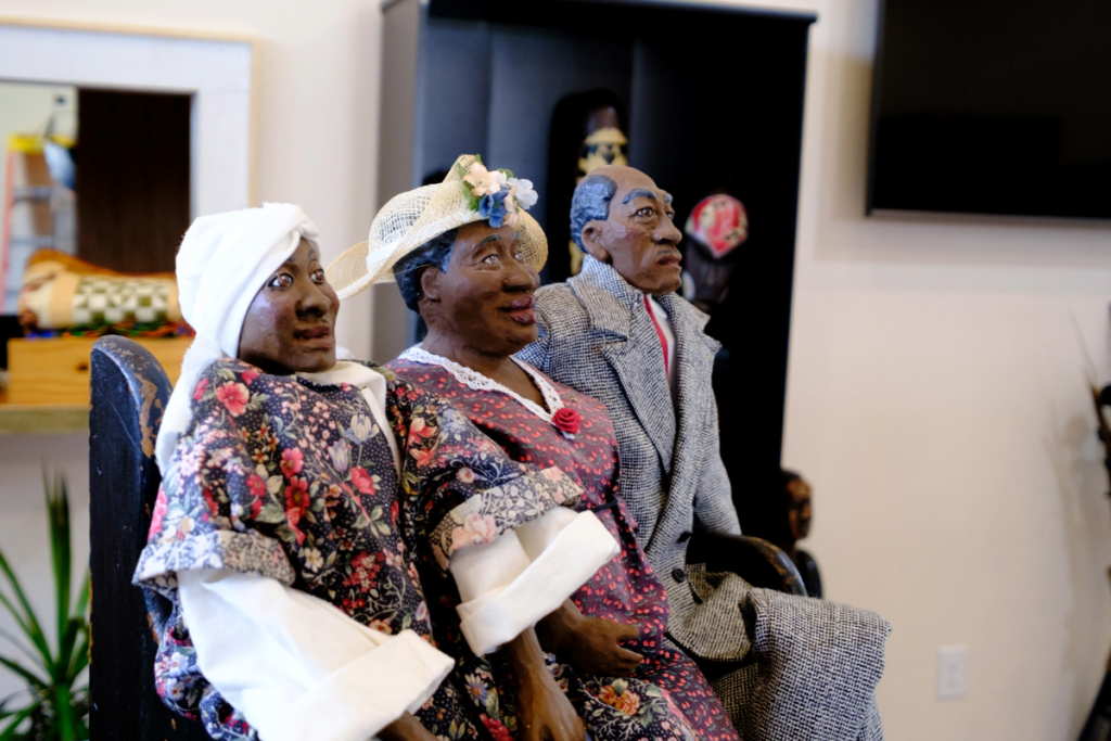 Three handmade dolls dressed in vintage clothing sit together on a bench at the Black History Museum. Two women wear floral dresses and hats, while the man wears a gray suit. The background features household items and artwork.