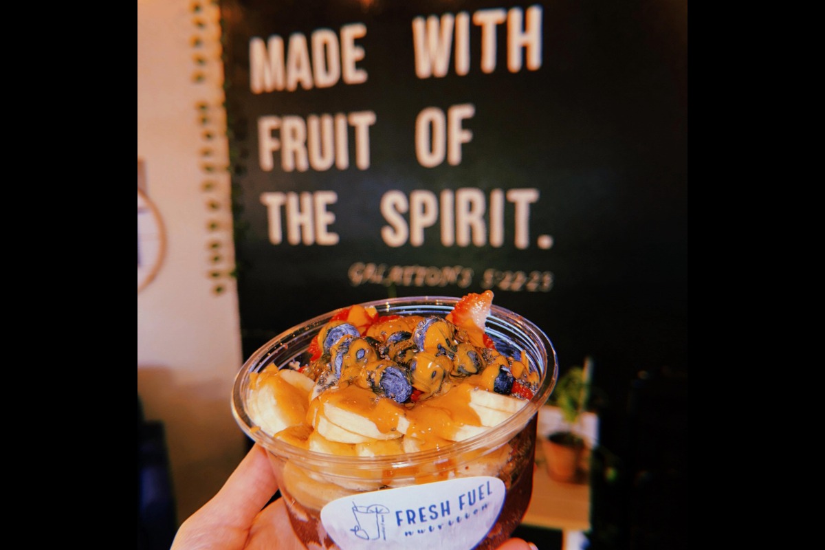 A hand holds a Fresh Fuel Nutrition bowl topped with sliced bananas, blueberries, strawberries, and nut butter. In the blurred background, a wall reads, MADE WITH FRUIT OF THE SPIRIT.