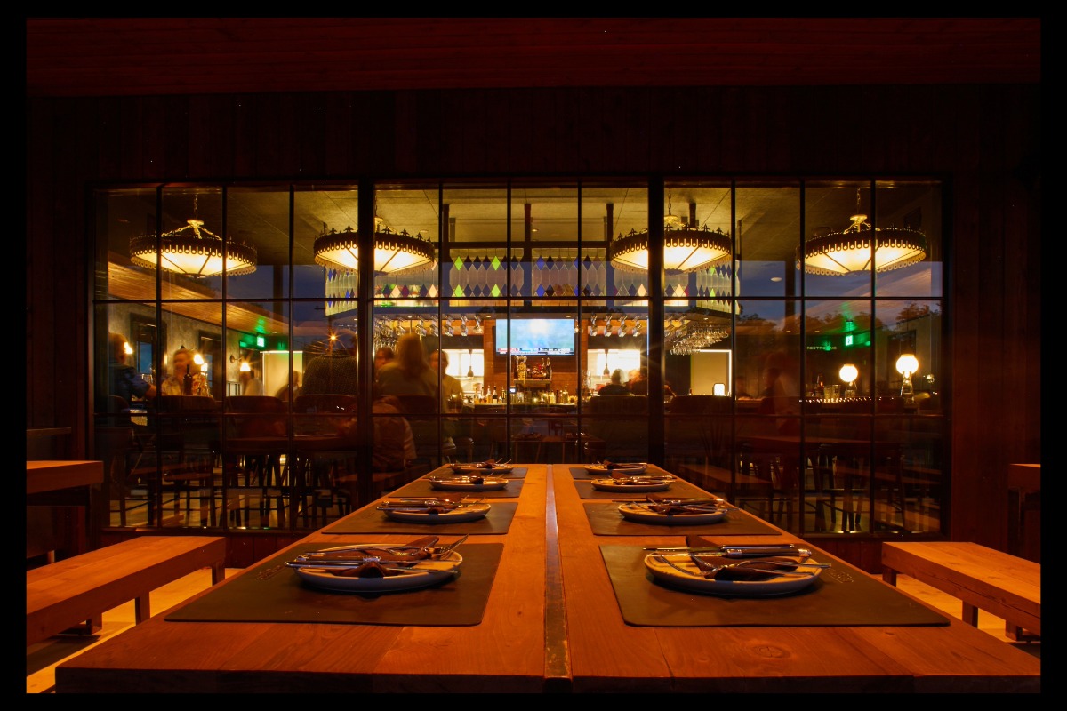 A warmly lit steakhouse interior viewed through glass walls, with long wooden tables set for fine dining in the foreground and a bar with glowing lights and people inside in the background at Junction Prime Steakhouse.