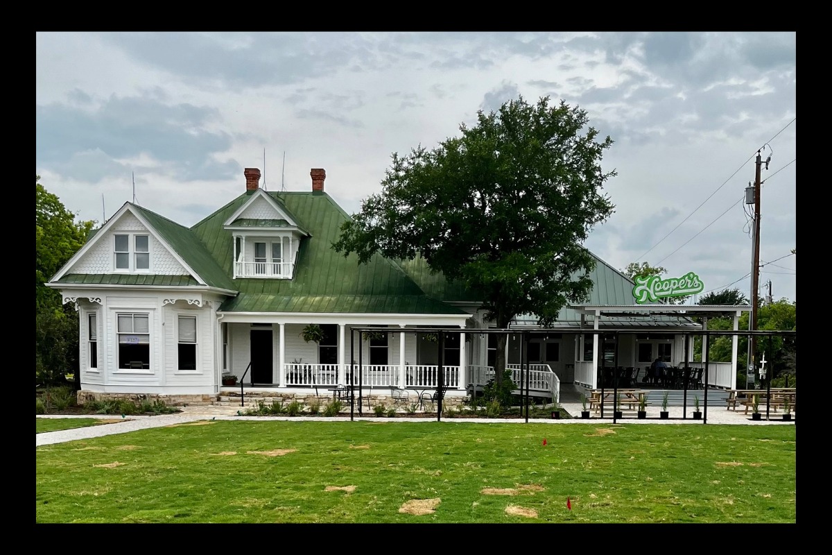 A large white house with a green roof and wraparound porch sits behind a lawn. An outdoor seating area is covered by a metal awning with a neon Hays Co. Oasis sign. Trees and a cloudy sky are in the background.