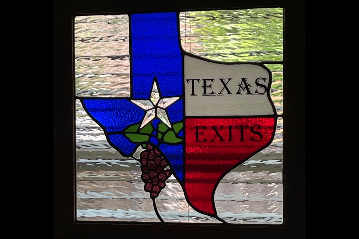 Decorative stained-glass window in the shape of Texas with blue, white, and red color blocks; a white star and grape cluster sit in the blue area, and the word 'TEXAS EXITS' appears in the panels on the right.