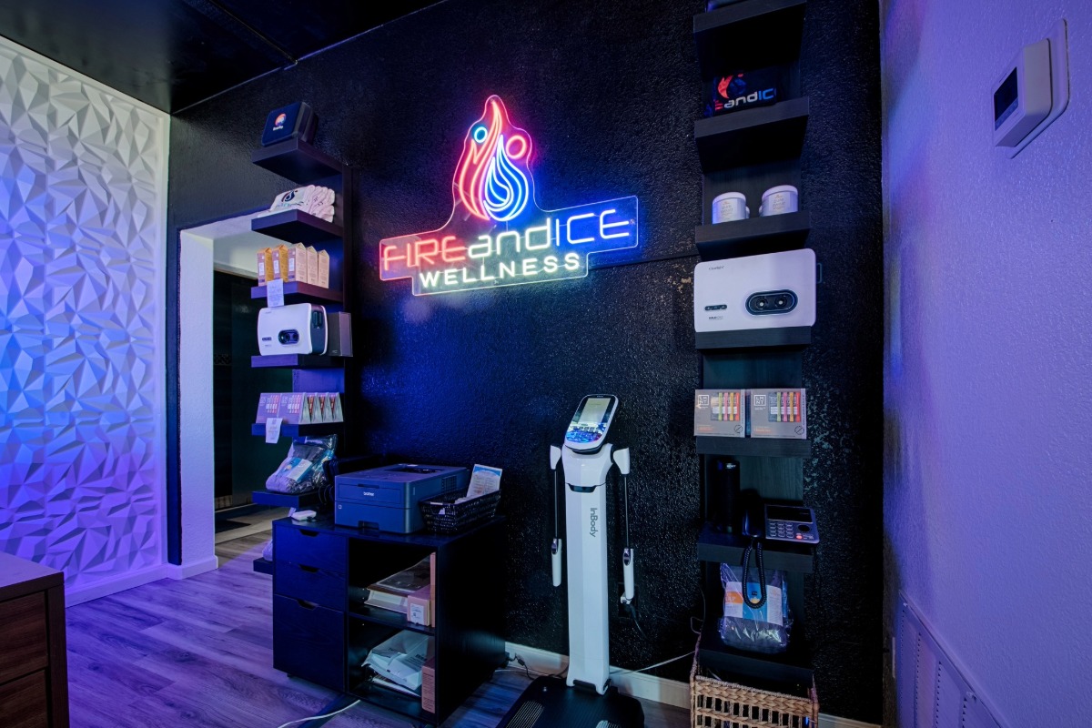 A modern wellness clinic interior with black walls, a neon Fire and Ice Wellness sign, shelves holding Fire and Ice Wellness products and equipment, a printer, digital scale, and a textured white accent wall.