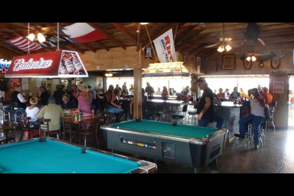 A lively scene at Reverend Jim's Dam Pub, where people sit at tables and the bar, surrounded by flags and beer signs. Two pool tables sit beneath a wooden ceiling with exposed beams and ceiling fans.