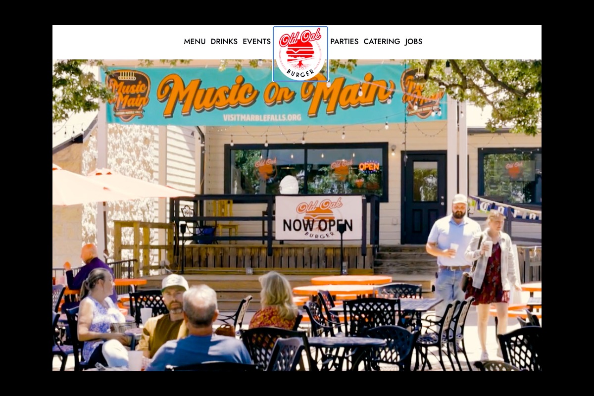 People sit and chat at outdoor tables in front of Old Oak Burger, with a large Music on Main banner and Now Open sign. Trees provide shade, and a few people are walking near the entrance.