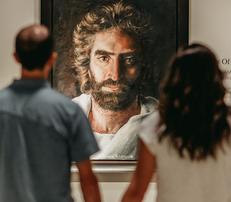 Two people stand hand in hand, viewed from behind, gazing at a realistic painting of a bearded man with long hair and striking eyes—one of Burnet County's unexpected finds—illuminated on a gallery wall.