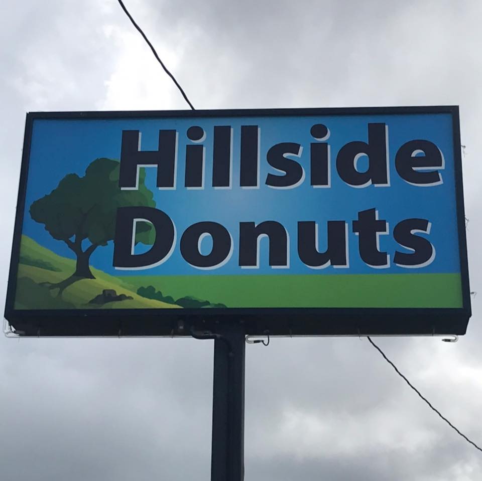A rectangular sign for Hillside Donuts bakery features bold black letters on a blue background with a green hillside, a tree illustration, and tempting donuts. Power lines and a cloudy sky are visible in the background.