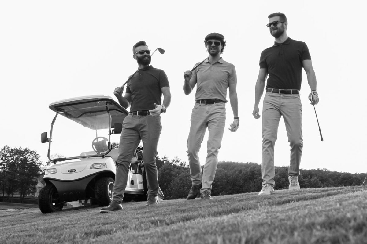 Three men in casual golf attire walk across a Burnet County golf course, smiling and holding clubs during their Guys Weekend. A golf cart and trees are visible in the black and white image.