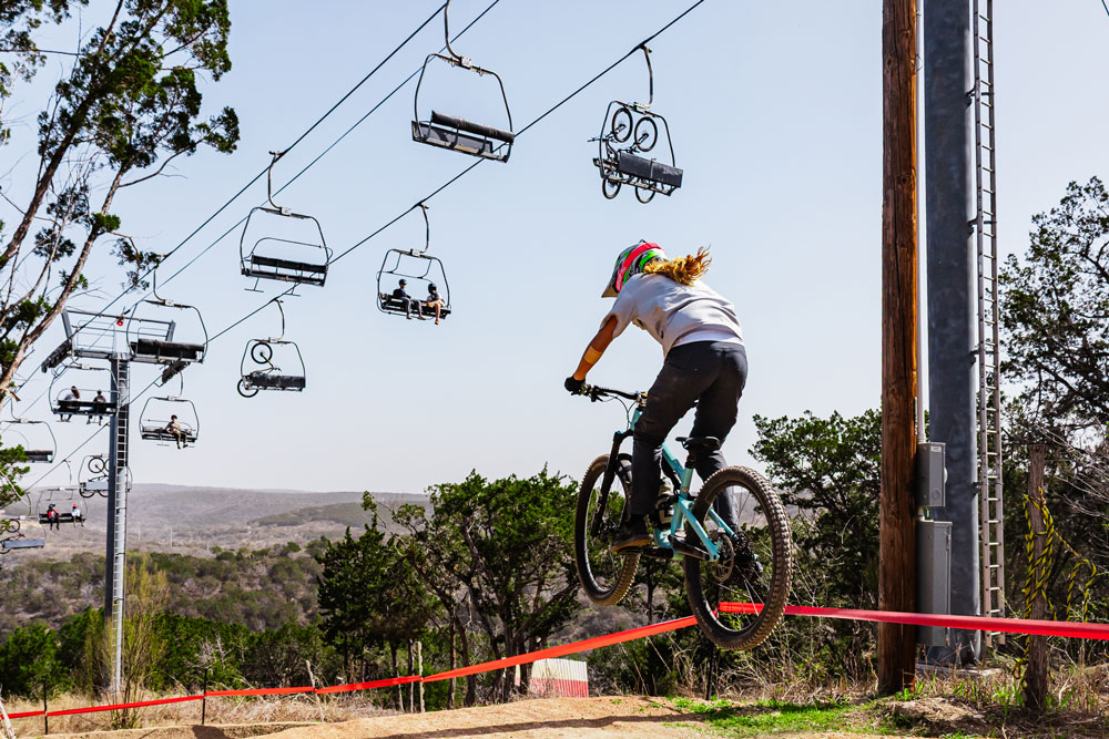 A mountain biker wearing a helmet jumps over a hill at Spider Mountain Bike Park, with ski lift chairs and cables overhead and a scenic landscape of trees and distant hills in the background.
