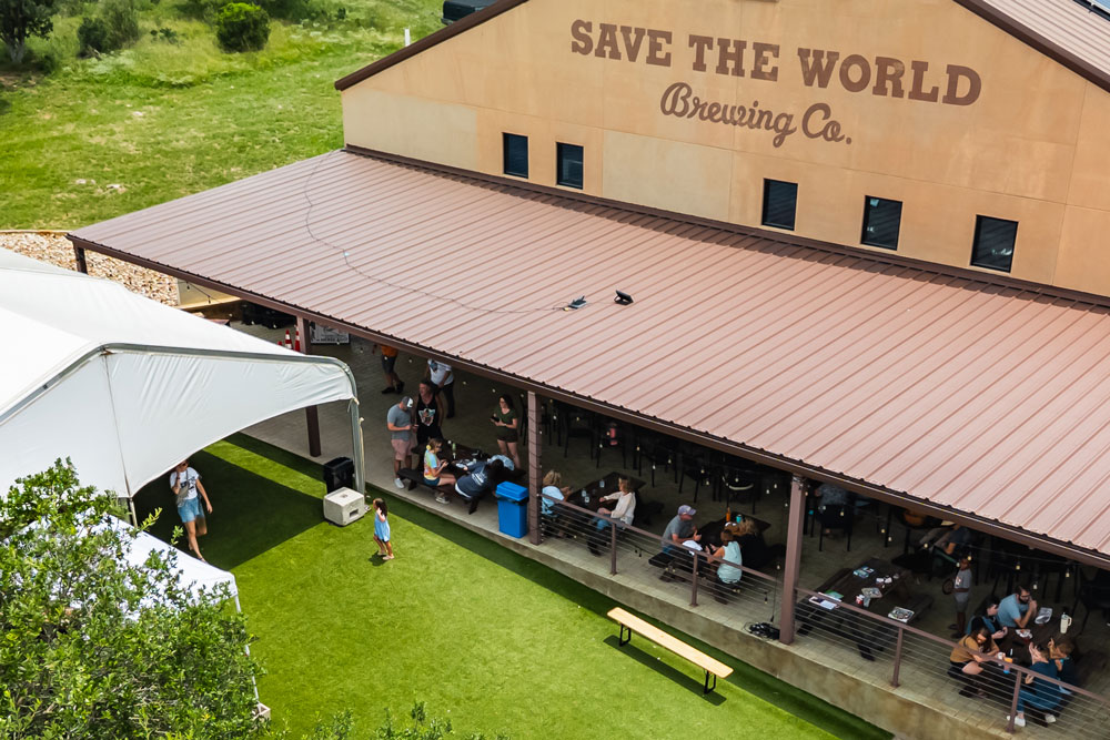 Aerial view of people socializing under a covered patio at Save The World Brewing Co. in Burnet County—perfect for a Guys Weekend or part of your 3 Day Itinerary. A grass lawn and large tent sit nearby, with the brewery’s name on the building.