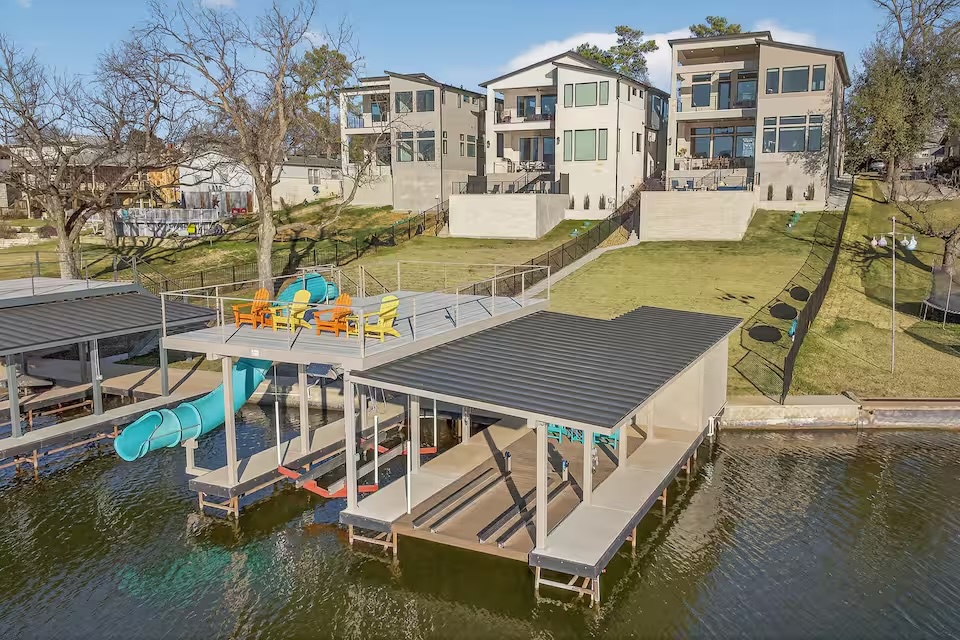 A modern Colorado Retreat lakeside property with two large homes overlooking the water, featuring a boat dock with covered slips, a slide, and colorful patio chairs on the dock’s rooftop deck. Grassy yard slopes down to the waterfront.