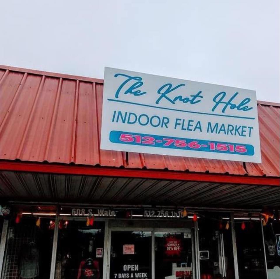 The exterior of The Knot Hole features a red roof and a large white sign with blue and red lettering above the entrance. The storefront windows display signs, including an "Open 7 Days a Week" notice, inviting shoppers to explore The Knot Hole's treasures.