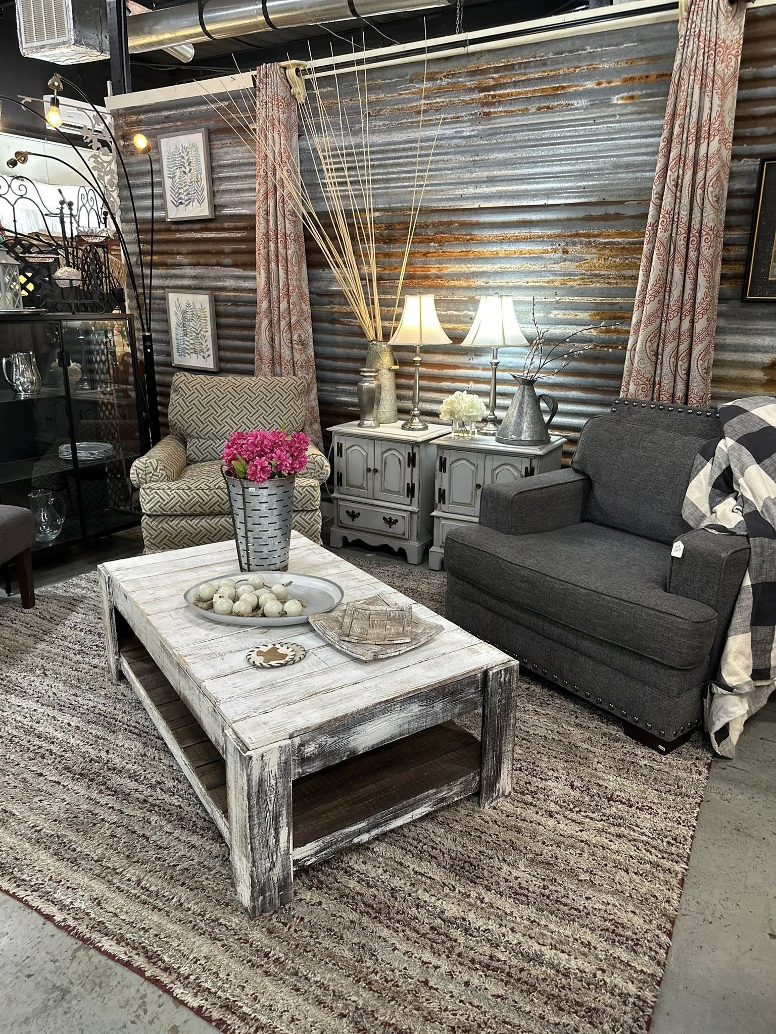A cozy living room with rustic decor from Pickins by Sassy features a whitewashed coffee table with a vase of pink flowers, two armchairs, a striped rug, side tables with lamps, and a corrugated metal wall backdrop.