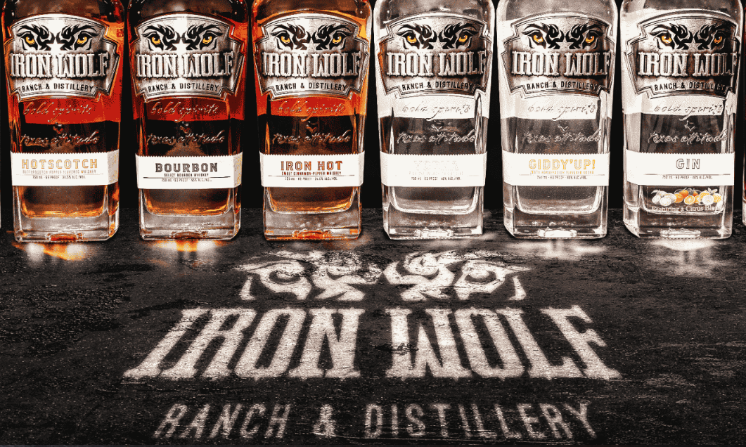 Six bottles of Iron Wolf spirits—Hotscotch, Bourbon, Iron Hot, two unclear labels, and Gin—stand in a row on a dark surface, with the Iron Wolf Ranch & Distillery logo from the renowned distillery projected below them.