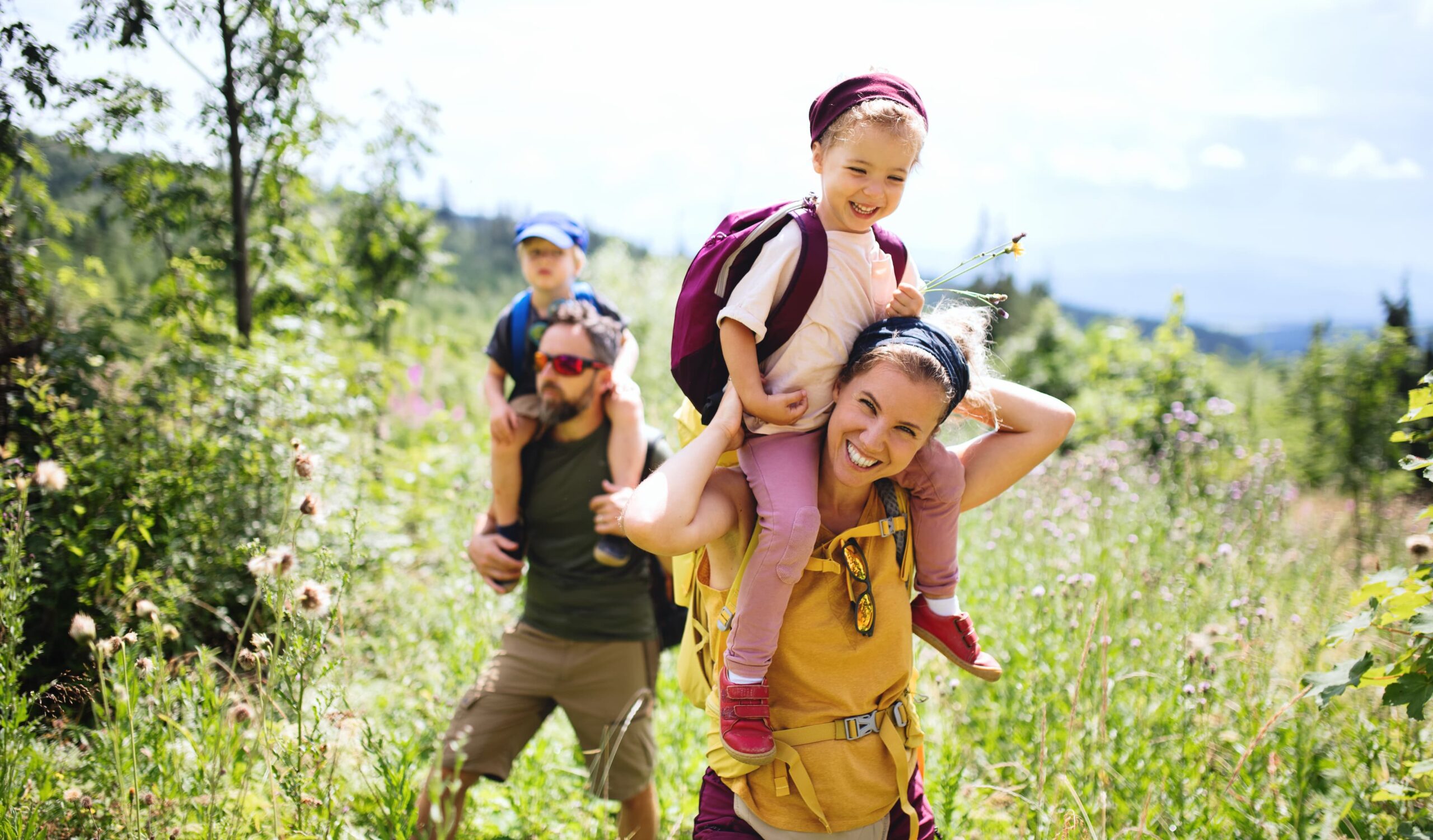 A smiling woman carries a young girl on her shoulders while hiking through a grassy, sunlit forest; behind them, a man carries a young boy on his shoulders. All appear happy and are wearing backpacks.