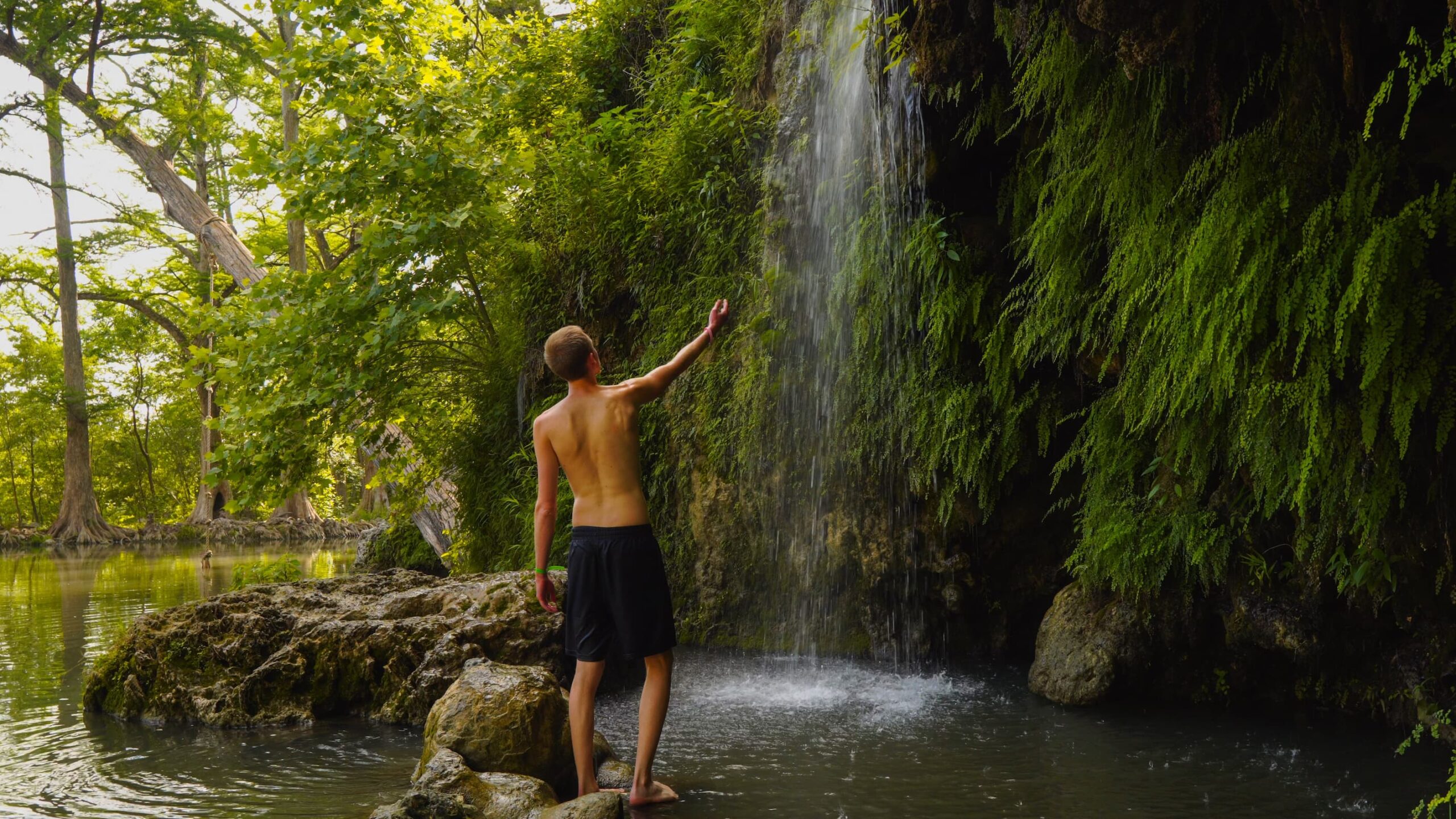 A person stands barefoot on rocks, reaching toward a small waterfall surrounded by lush green foliage in Burnet County’s Highland Lakes, with a calm river and trees in the background—perfect for a Family Weekend adventure.