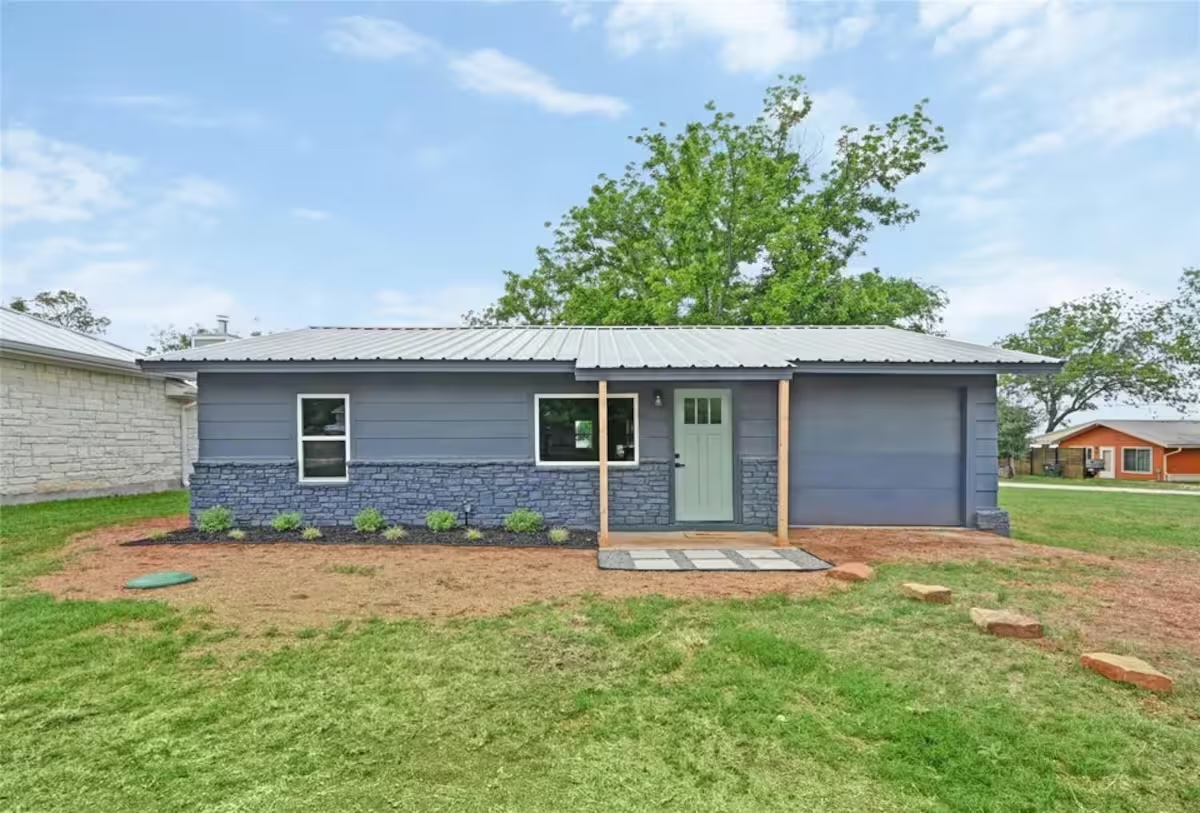 Single-story blue house with a light green door, gray metal roof, and stone accents. Small shrubs line the front, and a young tree stands behind the house. A stone path leads to the entrance, with grass and bare patches in the yard.