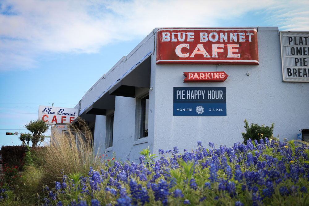 A café with red signs reading Blue Bonnet Café and Pie Happy Hour stands behind blooming bluebonnet flowers in Burnet County under a partly cloudy sky—a perfect stop on your 3 Day Itinerary or Guys Weekend adventure. A parking sign is also visible.