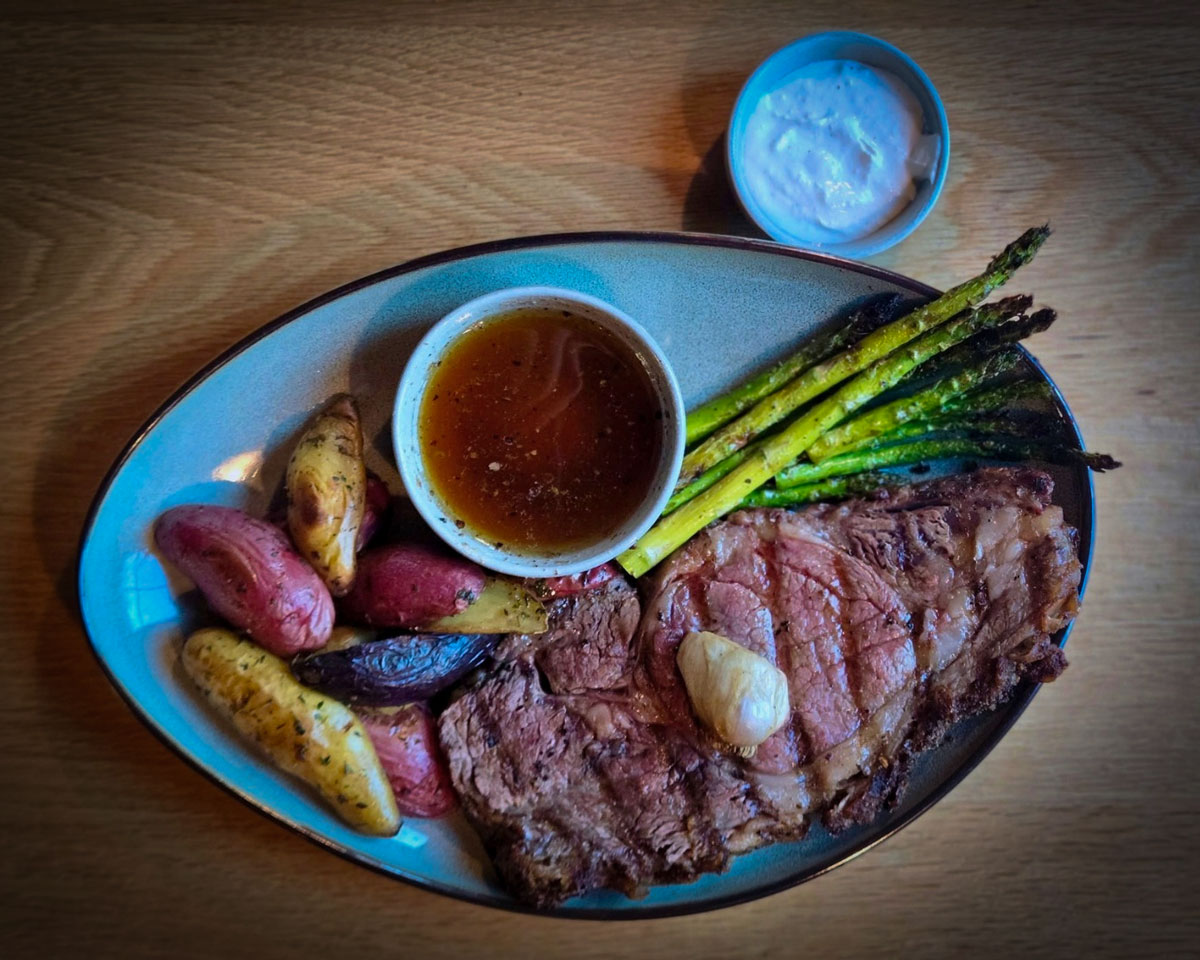 Perfect for a Guys Weekend in Burnet County, this plate features grilled steak with a dollop of butter, roasted fingerling potatoes, grilled asparagus, brown sauce, and a small cup of creamy white sauce on a wooden table.